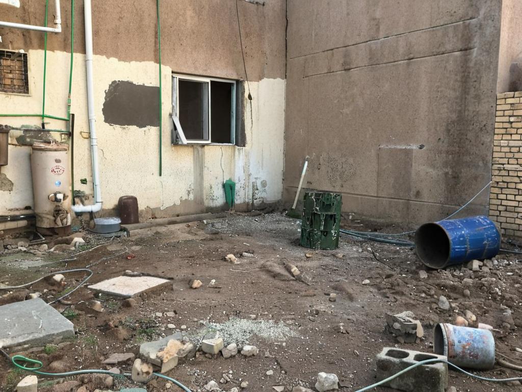 The photo presents an architectural outdoor garden scene in a state of obvious disrepair, which feels vintage. The walls are a mix of rough, textured plaster, painted in varying shades of off-white and tan, with visible imperfections adding to the aged aesthetic. A simple window is set into one of the walls, its frame appearing weathered. The ground is covered in dirt and debris, scattered with items like barrels (one blue, one black), bricks, and scraps of what appears to be construction materials. A complex network of pipes, painted green and white, runs along the walls, an old water heater is also visible, indicating a utilitarian space. The lighting seems to be natural, possibly taken during daylight, further emphasizing the roughness and unfinished look of the scene. This image might be considered vintage interior design, with a raw appeal that could make it trendy, and could be seen trending on pinterest.
