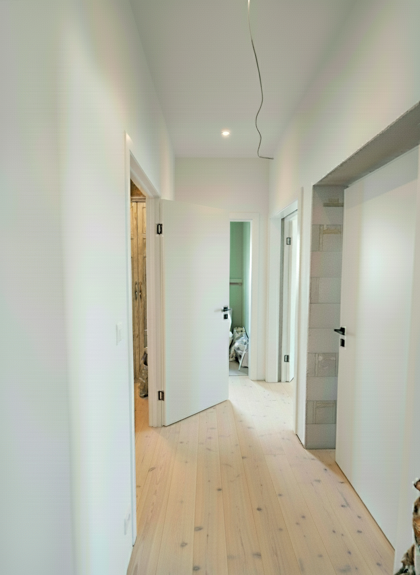An incredibly lifelike, richly detailed, photorealistic interior photo of a hallway leading to various rooms, set in a residential home under renovation. The scene is illuminated by natural midday light pouring in from unseen windows, creating a bright yet slightly unfinished atmosphere. Captured with a wide-angle lens, emphasizing concrete floors with paint splatters, raw cinderblock pillar details, and stark white walls, the image hints at a "shabby chic" aesthetic in its nascent stages, with the promise of warmth and eclectic charm.

Imagine this hallway transformed into a welcoming portal of understated elegance. The concrete floors are now polished and sealed, gleam enticingly under soft, recessed lighting, transitioning seamlessly into the rooms beyond. To the left, a doorway opens into a dressing room, meticulously organized with built-in wardrobes featuring distressed wood finishes and brushed brass hardware. The walls are painted a soft, muted sage green, creating a calming contrast to the bright white hallway. Delicate lace curtains hang from a window, catching the gentle breeze. To the right, the other doorway reveals a light-filled living space. Here, plush, overstuffed armchairs in faded floral patterns are clustered around a vintage-style coffee table. An antique Persian rug anchors the seating area, its worn fibers telling a story