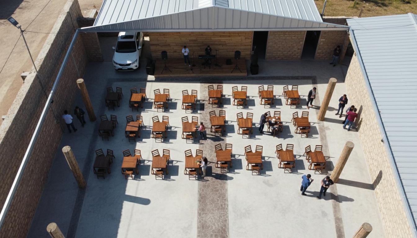 An incredibly lifelike, richly detailed, photorealistic aerial shot of a lively event space set in a modern fusion setting. The scene is illuminated by bright midday sun, creating a vibrant yet relaxed atmosphere suitable for an outdoor gathering. Captured with a wide-angle lens, emphasizing the expansive arrangement of tables and chairs, the polished concrete floor, and the rough-hewn taquique walls. The space is designed as a "Salon de fiestas," a venue for celebrations, with rows of square tables, each set with chairs, arranged on a light gray polished concrete floor. A contrasting band of stamped concrete defines a central pathway. A rustic wooden stage occupies one end, hosting a live band preparing for a performance, complete with a keyboard, microphones, and speakers. A white SUV is parked under a partially covered area, suggesting arrival. The building structure features prominent taquique walls and a corrugated metal roof supported by light gray steel beams. Thick wooden columns provide structural support at the edge of the main gathering area, hinting at an open-air but defined space. The overall impression is one of a functional, welcoming outdoor venue ready to host a festive occasion, bathed in the clear light of day.