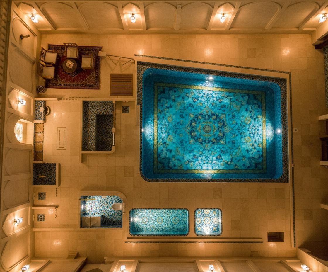 An incredibly lifelike, richly detailed, photorealistic photograph of a small swimming pool area at a traditional Iranian hotel. The scene is illuminated by warm, ambient light emanating from traditional Iranian wall sconces, creating an inviting and mystical atmosphere. Captured with a wide-angle lens, emphasizing the serene environment and intricate details, this shot showcases the main swimming pool occupying the upper right portion of the frame. The pool's surface shimmers, reflecting the soft light and hinting at the exquisite Iranian mosaic tile work that adorns its depths. In the lower section of the photograph, two smaller pools are visible, each with a gentle, inviting glow. The overall composition draws the viewer into the tranquil space, suggesting a deeply relaxing experience. The textures of the mosaic tiles, the smooth water surfaces, and the stylized illumination from the sconces are rendered with stunning clarity, transporting the viewer to this secluded oasis.
