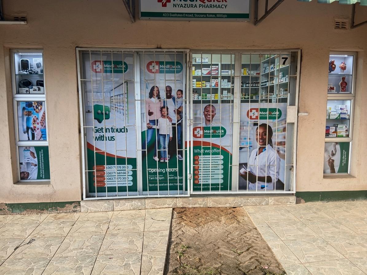 The photo captures the exterior of a pharmacy, "MediQuick Nyazura Pharmacy," presented in a straightforward, utilitarian architectural style. The building's facade is a simple, unadorned cream-colored stucco with a band of peeling green paint near the base. Large glass doors and windows dominate the storefront, covered by white metal security grills, suggesting a functional rather than purely aesthetic approach to design. The glass panels are extensively decorated with large, vibrant graphics advertising the pharmacy's services and branding. These decals, featuring images of families, smiling healthcare professionals, and various medical products, lend a somewhat busy, commercial feel that contrasts with the otherwise plain structure. A paved walkway, made of light-colored, irregularly patterned tiles, leads to the entrance. The lighting suggests a bright midday in what could be summer, with the sun casting subtle shadows, highlighting the textured surface of the tiles and the slightly uneven stucco of the building. The overall impression is one of practicality and accessibility, with the graphics serving to draw attention and inform passersby about the pharmacy's offerings.