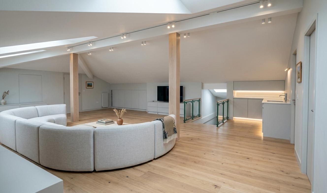 An incredibly lifelike, richly detailed, photorealistic interior photo of an attic loft, bathed in the soft, diffused light of midday. The scene is illuminated by a sophisticated track lighting system integrated into the white, vaulted ceiling, creating a bright and airy atmosphere. Captured with a wide-angle lens from a low perspective, emphasizing the spaciousness and the interplay of light and shadow, the rendering highlights the warm, light oak flooring that spans the open-plan living area. Dominating the foreground is a sleek, modern, light-colored curved sofa, its form a soft counterpoint to the clean lines of the architecture. To the right, a minimalist mini-kitchen with subtle under-cabinet lighting features light gray cabinetry and a contemporary sink, seamlessly integrated into the space. A striking green railing with glass panels and wooden handrails guides the eye towards a slightly elevated area, hinting at clever use of the attic's unique geometry. The walls, a soft, neutral light gray, enhance the minimalist aesthetic, contributing to the overall sense of tranquility and understated elegance.