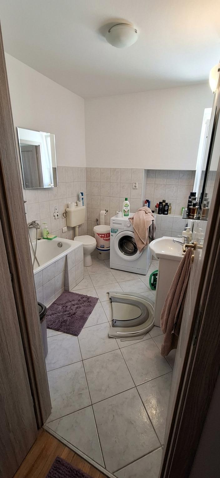 An interior photo of a bathroom, captured with a wide-angle lens to showcase the entire space. The overall style is contemporary elegance, exuding a clean, modern, and sophisticated ambiance. The walls are adorned with large, seamless porcelain slabs in a soft, creamy white, reflecting light and creating a sense of spaciousness. The floor is tiled with large format, high-gloss Carrara marble-effect porcelain tiles, lending a luxurious and polished feel.

The centerpiece of the bathroom is a freestanding soaking tub made of polished white resin, its smooth curves echoing the contemporary design. Adjacent to it, a wall-mounted vanity with a single, clean-lined basin is finished in a shimmering, pearlescent lacquer, its drawers featuring minimalist handles. Above the vanity, an oversized frameless de-fogging mirror reflects the room, doubling its perceived size. The lighting is a combination of recessed LED spotlights providing a cool, white general illumination, and a subtle, warm under-cabinet glow from the vanity, creating a spa-like atmosphere.

A sleek, wall-hung toilet with a concealed cistern is positioned discreetly in a corner, maintaining the room's uncluttered aesthetic. The shower enclosure is built with minimalist, frameless glass panels, featuring a large rainfall showerhead and a linear drain. Towel bars made of brushed nickel are strategically placed for convenience.

The color scheme is predominantly white and subtle monochromatic tones, accented by the natural veining of the marble-effect tiles and the subtle sheen of the pearlescent vanity. A single, large potted Fiddle Leaf Fig tree is placed in a corner, introducing a touch of organic texture and vibrant green to the serene environment.

The atmosphere is tranquil and serene, evoking a sense of calm and rejuvenation. The natural light is channeled through a large, frosted skylight overhead, diffusing a soft, indirect glow throughout the day. In the evening, the artificial lighting creates a warm and inviting sanctuary, perfect for relaxation. The overall design prioritizes function and form, with every element contributing to a harmonious and elegant experience.
