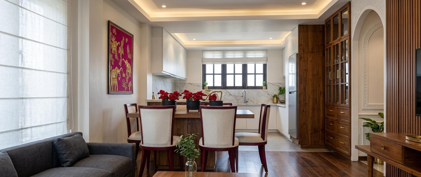 An incredibly lifelike, richly detailed, photorealistic wide-angle interior shot of a sophisticated open-plan living and dining suite set in a contemporary luxury residence. The scene is illuminated by soft, natural daylight diffused through elegant white Roman shades and warm recessed ceiling lights, creating a serene and opulent atmosphere. Captured with a high-end full-frame camera and a 35mm lens, emphasizing the intricate textures of handcrafted Indian textile wall art featuring golden elephants against a vibrant magenta background and the deep, lustrous grain of carved teak furniture. In the foreground, a charcoal linen sofa offers a plush textural contrast to the polished dark hardwood flooring. The dining area centers the space with ivory upholstered chairs trimmed in a rich, dark burgundy wood, surrounding a table adorned with striking red floral arrangements in artisanal black pots. To the right, a grand wooden hutch featuring brass-accented drawers and glass-paned doors stands next to a decorative arched wall niche, perfectly embodying the spiritual and ornate essence of the Indian Exuberance style. The transition into the kitchen reveals minimalist white cabinetry and a classic marble backsplash, framing the dark-mullioned windows and grounding the space with a modern touch, while scattered indoor greenery and handcrafted brass details add layers of warmth and organic vitality to this meticulously curated sanctuary.