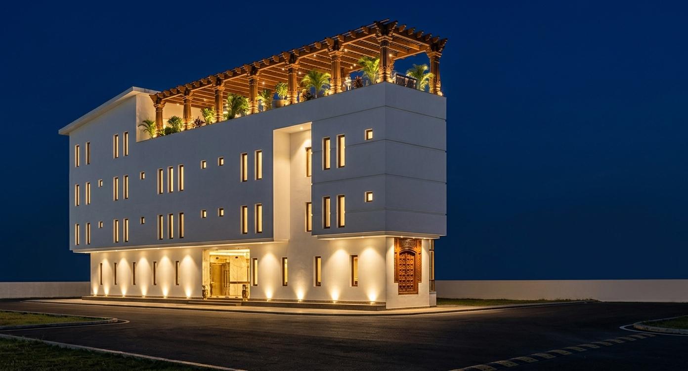 An incredibly lifelike, richly detailed, photorealistic wide-angle architectural shot of a majestic boutique hotel set in a serene urban environment during a balmy summer night. The scene is illuminated by a sophisticated network of warm recessed uplighters and the radiant golden glow emanating from the grand foyer, creating a welcoming and opulent atmosphere. Captured with a high-end 35mm tilt-shift lens, emphasizing the crisp geometric lines of the pristine white plaster facade and the intricate teak woodwork of the crowning pergola, the image showcases a masterpiece of modern design infused with Indian exuberance. The building features a striking cantilevered upper volume that hovers over a transparent ground-floor lobby, where polished marble and ornate brass accents are visible through the floor-to-ceiling glazing. A magnificent, hand-carved mahogany entrance door serves as a focal point, its intricate traditional motifs catching the light. Atop the structure, a vast rooftop terrace is defined by a rhythmic colonnade of decorative pillars supporting a sprawling wooden trellis, under which soft ambient lighting reveals a lush garden sanctuary. The deep indigo sky of the summer night provides a high-contrast backdrop, reflecting off the dark, polished asphalt of the approaching driveway and highlighting the building’s clean, rhythmic window placement and sophisticated architectural silhouette.
