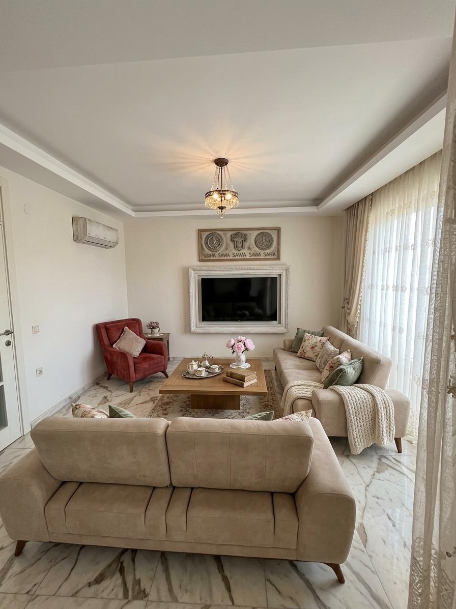 An incredibly lifelike, richly detailed, photorealistic wide-angle interior shot of a reimagined living room reimagined in a soft, shabby chic style set in a sun-drenched, airy home. The scene is illuminated by gentle, diffused afternoon light streaming through floor-to-ceiling lace and sheer linen curtains, creating a nostalgic and tranquil atmosphere. Captured with a high-end 35mm lens, emphasizing the intricate textures of embroidered textiles, weathered wood, and the subtle grain of the walls. The existing plush beige sofas are revitalized with an abundance of decorative pillows in faded floral prints and sage green velvet, while a chunky knit throw in cream drapes over one arm. The deep red velvet armchair provides a classic pop of color, now grounded by a large, antique-style rug with a worn, floral medallion pattern that softens the marble flooring. The bulky TV unit has been removed, leaving the space feeling expansive and organized, with a large flatscreen TV now mounted elegantly on the wall, encased in a custom-built, white-washed ornate wooden frame that transforms it into a piece of art. The square wooden coffee table is adorned with charming accessories: a silver tray holding a vintage tea set, a stack of linen-bound journals, and a small vase of soft pink peonies. Overhead, a reclaimed iron chandelier with delicate crystal drops replaces the old fixture, casting a warm glow that harmonizes the blend of rustic elegance and cozy, lived-in charm.