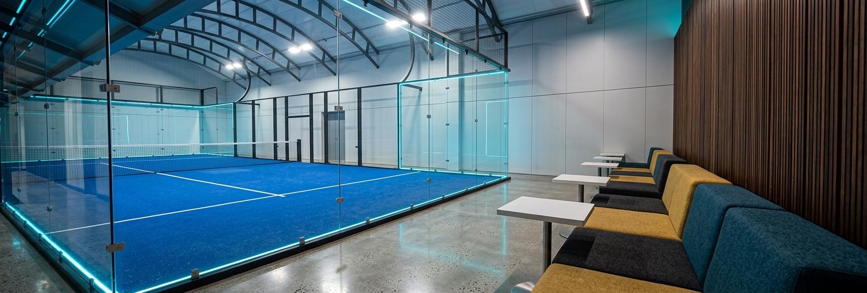 An incredibly lifelike, richly detailed, photorealistic wide-angle architectural photo of an indoor padel court arena and spectator exhibition space set in a cutting-edge athletic facility with a cyber eclectic fusion aesthetic. The scene is illuminated by crisp, high-intensity overhead stadium LEDs diffused through the vaulted structural steel ceiling, coupled with atmospheric neon-blue ambient glows reflecting off the glass court walls, creating a vibrant, high-energy, yet sophisticated atmosphere. Captured with a professional 24mm tilt-shift lens, emphasizing the crisp geometric lines and raw material textures, the view highlights a perfectly manicured blue synthetic turf court enclosed by massive frameless glass partitions on the left, which mirror the striking arched roof trusses above. To the right, a sleek spectator lounge features minimalist white-topped pedestal tables resting on polished concrete floors, adjacent to low-profile modular bench seating upholstered in tactile fabrics of dark charcoal, mustard yellow, and deep teal blue. Backing this comfortable viewing area is a striking feature wall of dark, warm vertical wooden slats, providing a tactile, organic contrast to the high-tech, athletic enclosure of the glass court.
