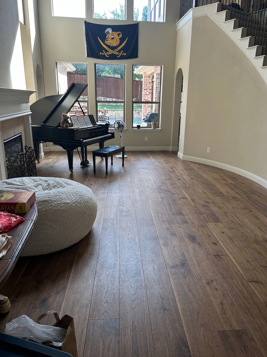 A professional photograph of the upscale living room, maintaining the exact same layout, geometry, camera angle, and composition, with all previous tiled and carpeted flooring replaced by a single, continuous expanse of premium wide-plank engineered hardwood flooring in a medium-dark warm wood tone. This new flooring showcases rich, organic grain patterns and a realistic satin finish that catches and softly diffuses the natural light. Bright daylight streams in through the large windows, casting long, soft-edged shadows across the new wood planks and highlighting their fine physical textures and seams. Every other element remains unchanged: the glossy black grand piano, the white textured bean bag chair, the fireplace, the beige walls, and the staircase on the right are all captured with absolute geometric fidelity. The scene is shot with a high-end DSLR camera, exhibiting natural depth of field, subtle lens vignetting, and authentic micro-imperfections like minor dust on the piano lid and soft reflections on the wood floor, giving the image a genuine, unstaged photographic quality.