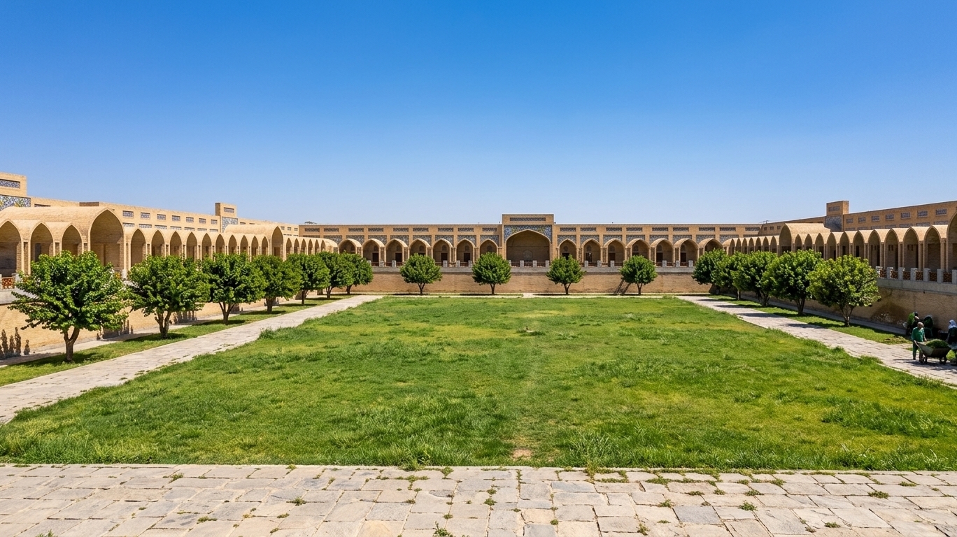 A high-resolution, professional wide-angle photograph of the expansive courtyard on a bright spring midday, captured under a clear, brilliant blue sky. The harsh, overhead sunlight casts sharp, short shadows directly beneath the architectural arches and the foliage, highlighting the warm, sandy-beige texture of the brick and plaster building facade. The building's intricate arches feature realistic Islamic-style glazed blue mosaic tiles that subtly catch the sun, and the walls show natural, subtle imperfections, faint weathering, and texture variations. The grand central lawn is a vibrant, lush spring green with realistic, individual blades of grass showing natural height and color variations. A well-defined stone path, made of weathered grey-paved slabs with rich, gritty textures in the seams, borders the lawn. The rows of manicured spring trees are dense with detailed green leaves, their bark having a rough, realistic texture, with the bright sunlight glinting off the upper canopy. The image has a deep depth of field, keeping the entire perimeter of the square in sharp focus, rendered with the natural micro-imperfections, faint atmospheric haze, and organic lighting of a genuine photograph.
