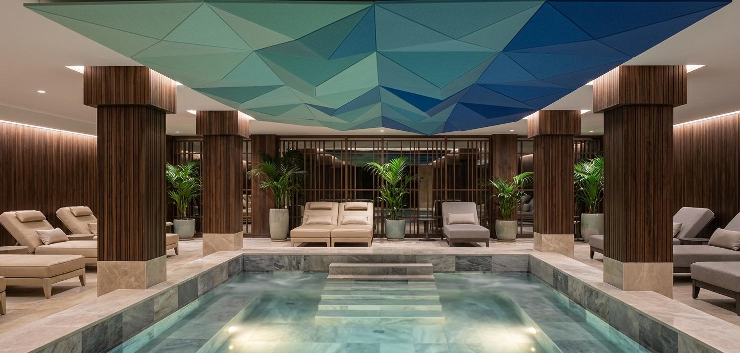 An incredibly lifelike, richly detailed, photorealistic wide-angle interior photo of a luxurious onsen and indoor wellness pool set in a contemporary high-end spa. The scene is illuminated by soft, integrated LED architectural lighting and a warm underwater glow, creating a serene, meditative, and sophisticated atmosphere. Captured with a professional 24mm tilt-shift lens, emphasizing the fine textures of polished stone, warm wood grains, and the fluid clarity of the water.

In the foreground, the pristine, steaming mineral water of the onsen pool sits beautifully within its basin, clad in variegated slate and polished travertine tiles of soft gray and pearl. A wide, symmetrical set of tiled steps with slim, dark metal handrails descends smoothly into the water, inviting relaxation.

Suspended overhead, the commanding geometric ceiling installation is reimagined as a stunning sculptural centerpiece. It features faceted, matte-finished custom acoustic panels composed of interlocking triangular planes in a gradient of seafoam green, teal, and deep cobalt blue, evoking the serene shifts of ocean depths. 

Flanking the pool, massive square columns are clad in deeply textured, dark vertical-grain walnut wood, resting on polished champagne-colored stone bases. On the surrounding decks, the lounging areas are populated by tailored, upholstered chaise lounges in soft champagne and neutral gray fabrics, arranged neatly for quiet contemplation. In the background, minimalist wooden screens and curated exotic potted palms in sleek ceramic planters add organic elegance to the clean-lined architecture. The overall composition is airy, balanced, and exceptionally refined, presenting a harmonious blend of Japanese-inspired minimalist wellness and contemporary luxury.