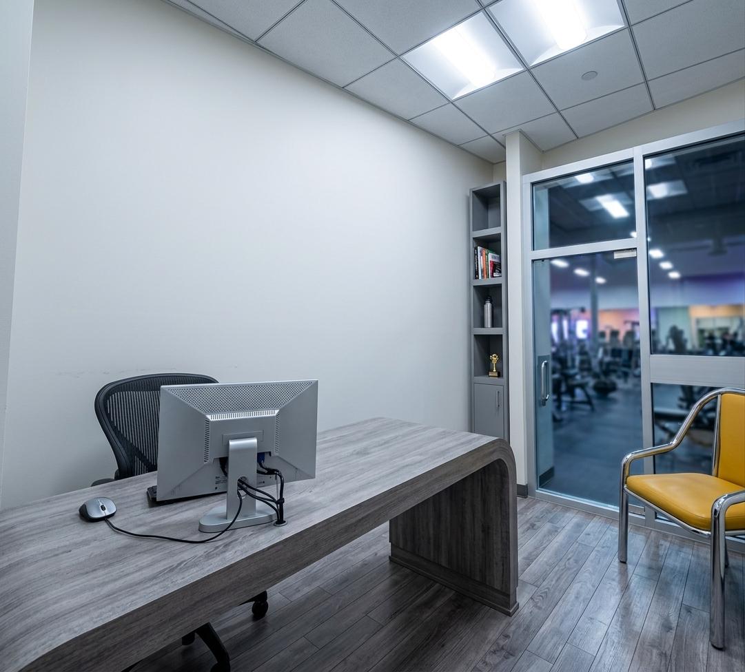 An incredibly lifelike, richly detailed, photorealistic wide-angle interior shot of a head coach's private office set in a modern fusion fitness gym. The scene is illuminated by cool-toned, recessed LED light panels in the crisp white grid ceiling and a vibrant ambient glow spilling from the adjacent workout floor, creating a highly professional and motivating atmosphere. Captured with a 24mm wide-angle prime lens, emphasizing the textured grain of the gray-washed parquet flooring and the matte surfaces of the contemporary office furniture, the image showcases a spacious, sleek workplace. In the foreground, a large composite executive desk with a curved gray frame anchors the room, supporting a modern, silver-backed flat-screen monitor with clean cable management and a wireless desktop mouse on its clean surface. Tucked neatly behind the desk is a black ergonomic office chair, while to the right sits a minimalist visitor's chair crafted with a lustrous tubular metal frame and a bright, mustard-yellow padded seat cushion. The right side of the frame features a floor-to-ceiling glass wall and electronic sliding door partition that serves as the entrance from the active main hall of the fitness gym, revealing out-of-focus silhouettes of exercise equipment in the background. Abutting this glass wall is a tall, streamlined gray cabinet featuring open display shelves on the upper section and solid, clean-cut cabinet doors on the lower half. The minimalist white walls and the clean lines of the design reinforce a state-of-the-art athletic-administrative aesthetic.