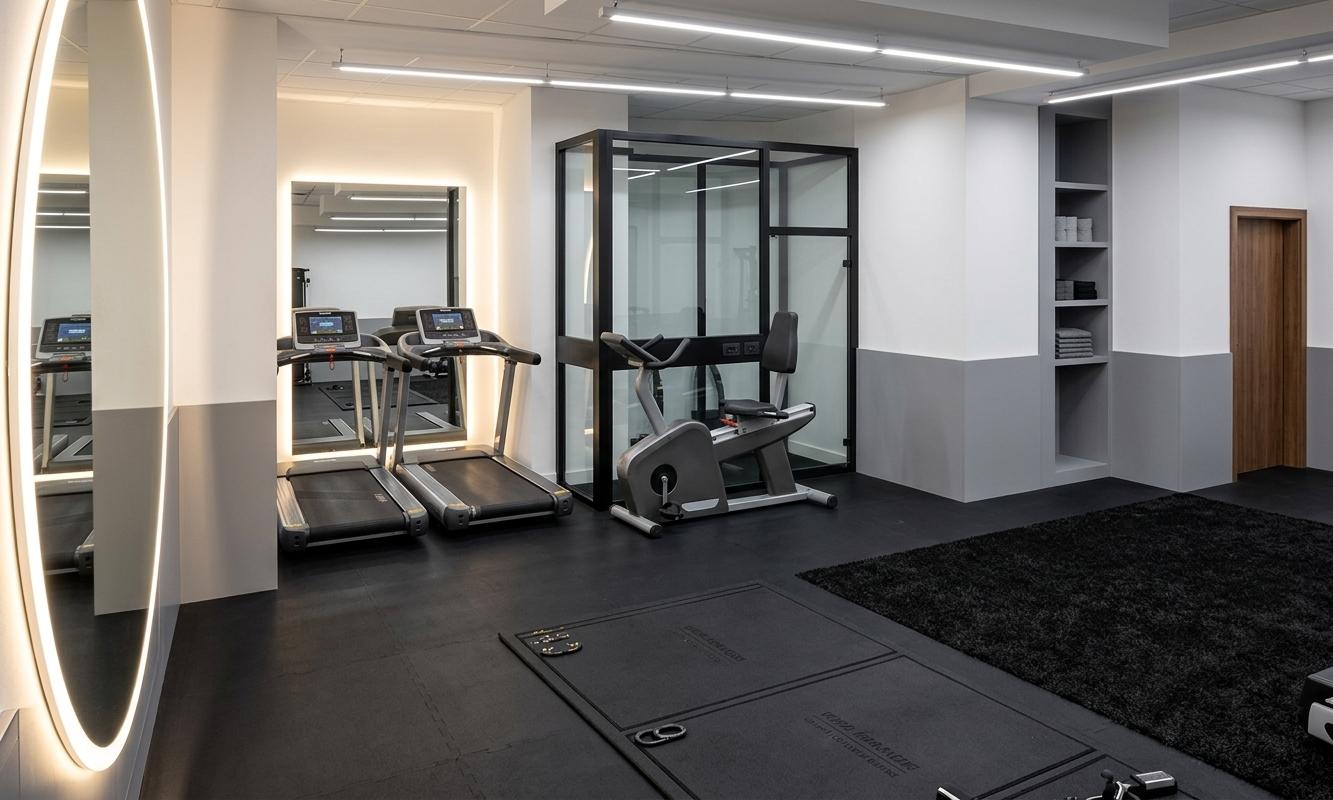 An incredibly lifelike, richly detailed, photorealistic wide-angle interior shot of a modern fusion fitness gym set in a high-end urban athletic club. The scene is illuminated by bright white linear LED track lights recessed into the clean white ceiling and a warm, glowing halo from a backlit mirror, creating an energetic, sleek, and highly sophisticated atmosphere. Captured with a professional 24mm wide-angle lens, emphasizing the tactile textures of the matte-black surfaces and the crisp, clean architectural finishes, the composition reveals an entirely black floor made of high-density rubber tiling integrated with a designated zone of textured black fake grass. The walls are a pristine, minimalist white, offset by smooth mid-gray wainscoting running along the lower perimeter, while the left wall features a massive, floor-to-ceiling mirror seamlessly framed by a brilliant white ring of diffused LED light. Modern, high-tech fitness equipment, including two sleek, silver-accented treadmills and a stationary exercise bike adjacent to a black-framed glass-partitioned booth, are precisely arranged in the space. In the background, a built-in recessed gray shelving unit and a warm wooden-door frame add functional sophistication, making this refined, monochromatic workout sanctuary feel incredibly tactile, spacious, and inviting.