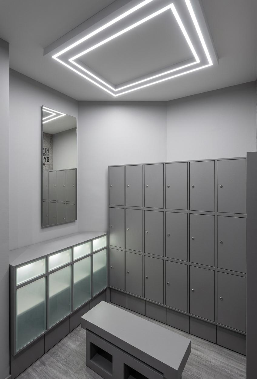 An incredibly lifelike, richly detailed, photorealistic wide-angle interior shot of a sleek changing room set in a modern fusion fitness gym. The scene is illuminated by crisp, cool-white linear LED strip lights beautifully recessed around a suspended white ceiling panel, creating a clean, sophisticated, and energetic atmosphere. Captured with a 24mm wide-angle lens, emphasizing the seamless, textureless matte gray MDF finish of the lockers and the delicate grain of the premium gray parquet flooring. On the right, a tall, multi-tiered grid of smooth gray lockers stands in sharp alignment, while on the left wall, a shorter single-tier row of matte gray closets with glowing frosted panels is positioned beneath a tall, crystal-clear vertical mirror that reflects the room's minimalist design. Centered on the elegant wood flooring is a solid, clean-lined gray bench with open cubbies, completing this highly-functional, pristine, and modern athletic wellness space.