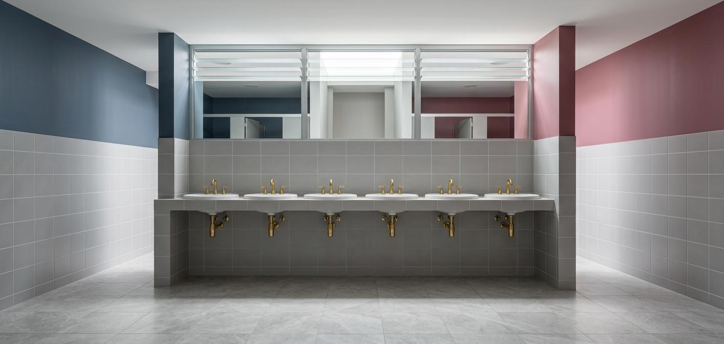 An incredibly lifelike, richly detailed, photorealistic interior photo of a contemporary, elegant washroom vanity area set in a modern boutique commercial space during a bright summer midday. The scene is illuminated by crisp, diffused sunlight filtering through high louvered glass transoms, creating a clean, serene, and highly sophisticated atmosphere. Captured with a professional 24mm architectural lens, emphasizing the glittering reflection on gold metallic fixtures and the pristine, polished textures of the ceramic tiling, the composition features a perfectly symmetrical, level perspective of a long floating vanity block. This central structure is wrapped in flawless, matte light-grey square tiles and holds six sparkling white undermount porcelain washbasins, each fitted with a brilliant, polished brass gooseneck faucet that glitters under the ambient light. The flanking walls showcase a sleek dual-tone design, transitioning from clean grey tiles on the lower half to smooth, matte-painted plaster above—with a deep slate-blue on the left and a refined, dusty berry-pink on the right. Above the main tiled partition, high-level horizontal louvers reveal glimpses of clean, minimalist stall compartments behind, enhancing the deep spatial perspective of this impeccably designed, luxury modern restroom.