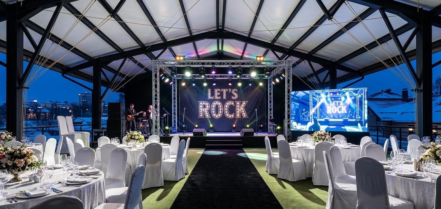 An incredibly lifelike, richly detailed, photorealistic wide-angle interior architectural photo of a lavish, contemporary gala event set in an expansive, attic-inspired rooftop pavilion during a serene winter night. The scene is illuminated by dramatic, multi-colored stage lighting and crisp spot beams reflecting off glittering table settings, creating a sophisticated and highly energetic atmosphere. Captured with a professional 24mm tilt-shift lens, emphasizing the sharp lines of the matte-black structural trusses and the clean, pristine surfaces of the white-clad furniture.

The overhead structure showcases a stunning contemporary attic design, featuring a soaring gabled roof supported by heavy, geometric black steel framing. Slender yellow tension cables cross beneath clean white canopy panels, creating an intricate geometric pattern overhead. Beyond the open sides of the pavilion, the cool, dark blue tones of a crisp winter evening create a stark, beautiful contrast with the warm, vibrant energy inside.

At the center of the space is a magnificent, rock-themed stage featuring a dark, professionally backlit banner that proudly displays "LET'S ROCK." The stage is framed by sleek metal lighting trusses loaded with state-of-the-art stage fixtures. To the left, performers stand near modern audio monitors and microphone stands, while to the right, a massive, ultra-thin widescreen monitor displays a brilliant, high-definition live feed of the performance, its glow casting realistic reflections on nearby surfaces. 

The dining area is the epitome of contemporary elegance, styled with clean, modern, and glittering white furniture. Numerous circular dining tables are meticulously arranged on a soft, moss-green carpeted floor, divided by a prominent, plush black aisle runner that leads directly to the stage. Each table is draped in immaculate white linens and surrounded by high-backed chairs dressed in form-fitting white slipcovers. Tabletop details sparkle in the light, featuring shimmering crystal glassware, polished silver cutlery, and elegant floral centerpieces with dark, velvety flowers. The dramatic overhead spotlights cast sharp, graphic shadows from the chairs onto the floor, enhancing the depth and spatial complexity of this luxurious setting.