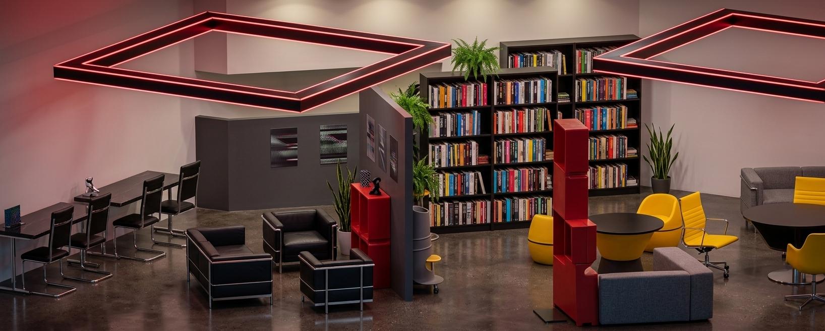 An incredibly lifelike, richly detailed, photorealistic wide-angle interior photo of a collaborative study room set in a vibrant techno wonderland. The scene is illuminated by a mix of soft, recessed ambient lighting and glowing crimson geometric overhead frames, creating an immersive, energetic, and futuristic atmosphere. Captured with a 24mm wide-angle lens, emphasizing the high-contrast textures of modular metal and polished plastic furniture alongside sleek, eclectic surfaces. Tall metal bookshelves packed with colorful photography books rise behind a central dividing wall, offset by darkaccent walls showcasing abstract glitch art wall art. To the left, a long study bar with tall chairs lines the edge, leading to a conversation nook composed of minimalist black leather armchairs with polished chrome frames on a smooth, industrial floor. On the right, a dynamic study hub features a circular workspace surrounded by striking red modular plastic panels, bright yellow accent chairs, and playful, techno-inspired decor. Touches of minimalist indoor plants are scattered throughout, injecting an energetic splash of green that balances the digital, high-tech aesthetic.