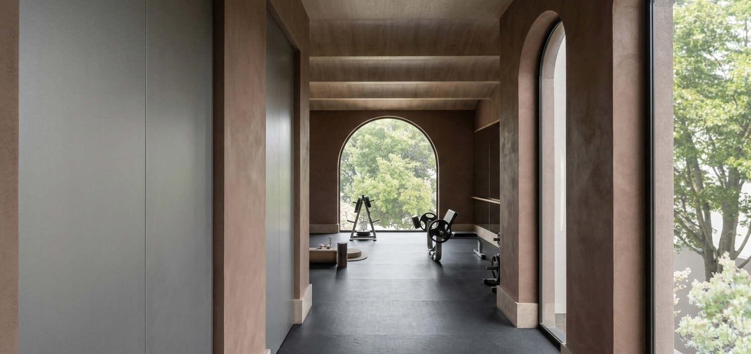 A high-end architectural photograph of the gym interior, preserving the exact layout, geometry, materials, and colors of the scene. The image is captured with a professional wide-angle lens, featuring soft, indirect daylight filtering through the large arched window at the far end and from the side openings. This diffused natural light gently illuminates the textured earthy-brown plaster walls and the light-colored wooden ceiling panels, casting subtle, realistic bounce light and soft gradient shadows along the surfaces. The dark, matte gym floor has a realistic rubberized texture that catches the light naturally, while the metal and rubber surfaces of the gym equipment show authentic, muted reflections and soft contact shadows on the floor. The exterior trees are visible through the window with a realistic depth of field, bathed in soft, overcast daylight. The composition and materials remain completely unchanged, rendered with photorealistic clarity, delicate ambient occlusion in the corners, and an authentic, high-quality photographic finish with subtle natural grain.