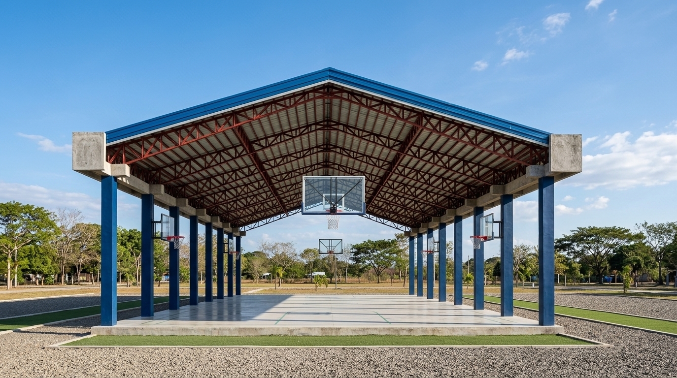An incredibly lifelike, richly detailed, photorealistic wide-angle architectural photo of an open-air basketball court and minimalist fitness gym pavilion set in a sun-drenched, serene outdoor recreational park. The scene is illuminated by crisp, natural midday sunlight that casts sharp, defined shadows across the polished concrete floor, creating a bright, energetic, and clean atmosphere. Captured with a professional 24mm tilt-shift lens, emphasizing the impeccably clean lines of the structural columns, the texture of the smooth concrete slab, and the vivid color contrast between the deep blue painted steel support pillars, the intricate red-painted overhead roof trusses, and the bright blue corrugated metal gabled roof. At each end of this industrial-inspired minimalist haven, professional-grade basketball hoops feature thick glass backboards, solid steel red rims with crisp white nets, and heavy-duty arm mounts anchored directly into the structure to complete the functional fitness courtyard. The concrete court is pristine, framed by a manicured gravel perimeter that transitions into low-profile green turf, while the background reveals a vast, clear blue sky decorated with soft, wispy cumulus clouds, creating a perfect harmony of architectural utility and modern athletic design.