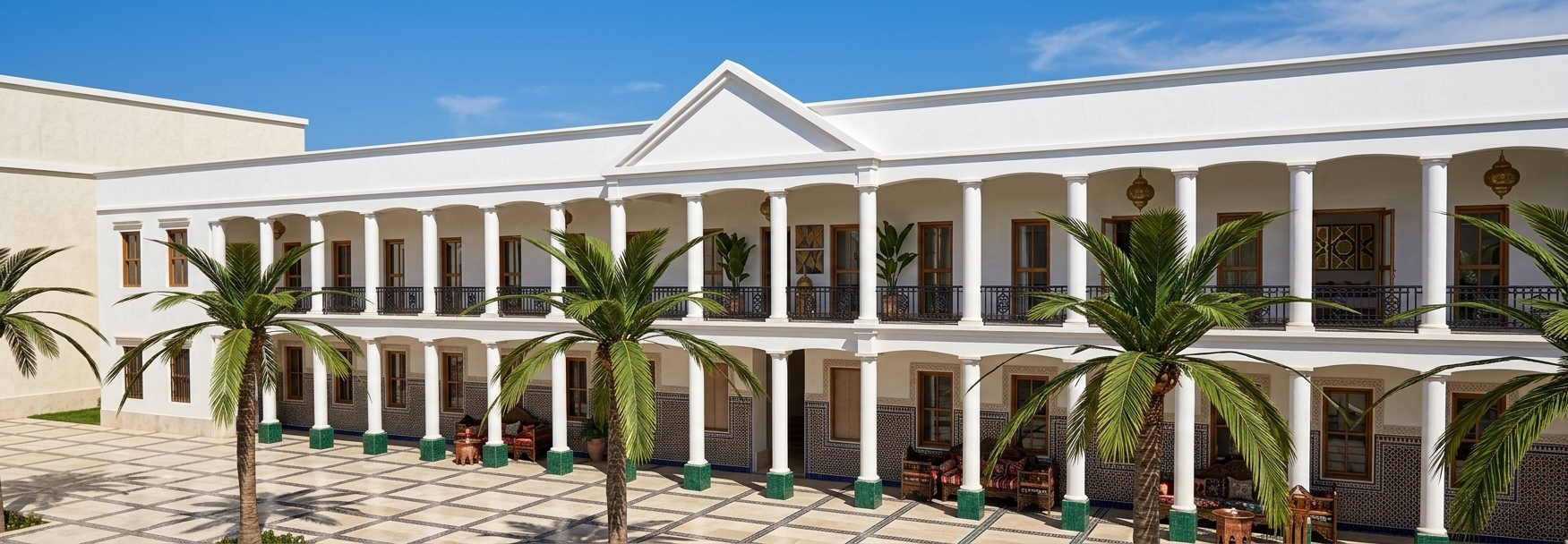 An incredibly lifelike, richly detailed, photorealistic wide-angle architectural photo of a grand two-story Moroccan-inspired pavilion set in a sun-drenched outdoor patio during a vibrant summer. The scene is illuminated by intense, direct midday sunlight, creating a warm, exotic, and sensual atmosphere. Captured with a professional medium-format camera and a 24mm wide-angle lens, emphasizing the rich textures of hand-carved wood, detailed metalwork, and intricate patterned surfaces, the architecture beautifully merges classical colonnades with Middle Eastern mystique. The building's lower level features a continuous gallery of pristine white columns resting on deep emerald-green bases, set against walls adorned with exquisite zellige tiles in geometric patterns of deep blues, dark reds, and shimmering golds. Along the expansive stone-paved patio, several pockets of lush, mature palm trees cast crisp, dramatic shadows, while ornate carved wooden and iron patio furniture with rich, colorful Moroccan textiles invite relaxation. The upper level showcases a brilliant white plaster facade punctuated by a central triangular pediment and a continuous balcony wrapped in an ornate, matte-black wrought-iron railing. Tall, elegant windows hint at luxury interior spaces decorated with geometric wall art, indoor plants, and traditional Middle Eastern brass lamps, presenting an overall composition that feels like a polished, vivid spread from a luxury travel and photography book.
