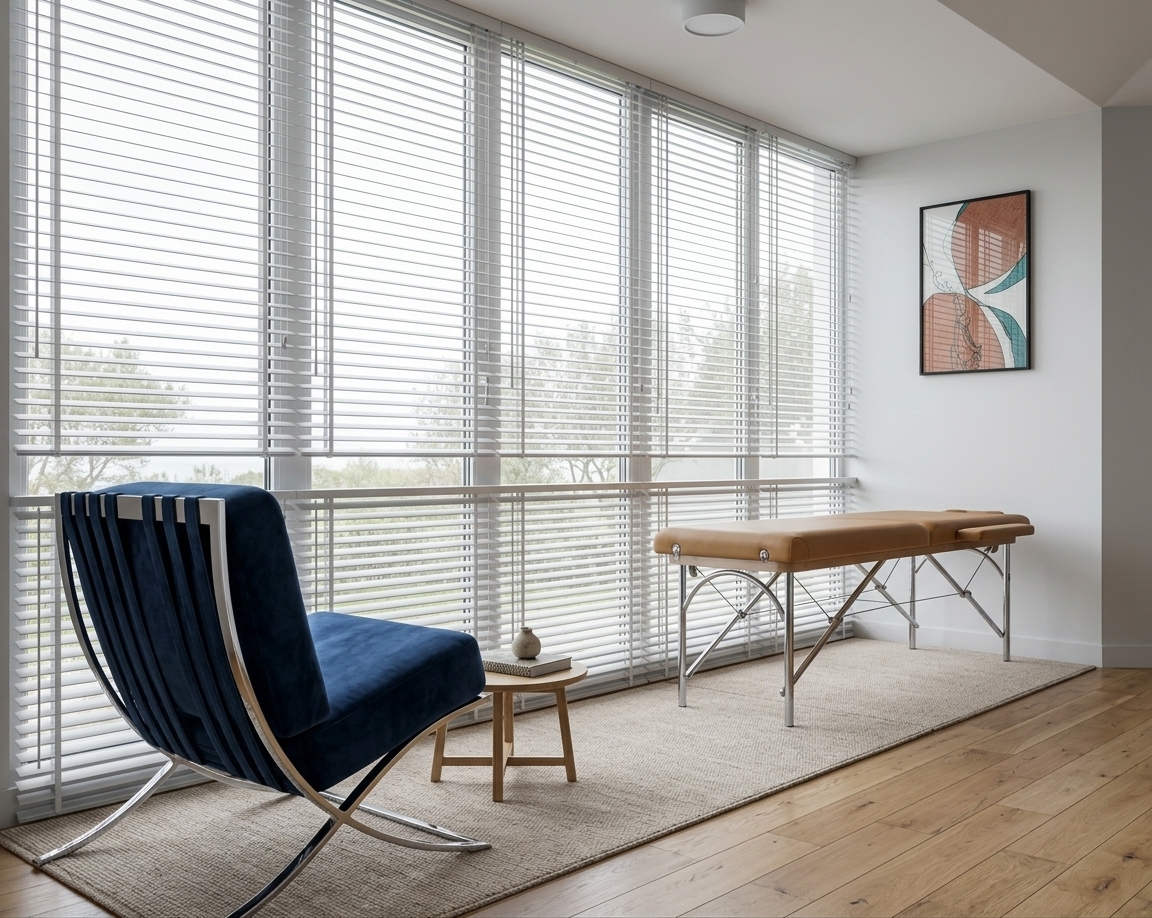 An incredibly lifelike, richly detailed, photorealistic wide-angle interior photo of a minimalist haven attic wellness space set in a tranquil coastal residence. The scene is illuminated by bright, diffused midday daylight filtering through expansive floor-to-ceiling glass windows with slim white horizontal blinds, creating a serene and Zen-inspired atmosphere. Captured with a professional 35mm lens, emphasizing the tactile textures of the plush navy blue velvet lounge chair with its sleek, curved chrome frame and the soft, woven beige area rug resting on the warm wooden floorboards. The room exhibits an uncluttered, modern aesthetic defined by clean lines and neutral shades of white and gray. A functional, streamlined folding therapy table with a tan upholstered top is positioned elegantly on the right, balanced by a piece of abstract wall art in terracotta and teal hues on the crisp white wall above it. A small, three-legged wooden side table occupies the middle ground, styled minimally with a photography book, enhancing the space's peaceful, contemporary allure.