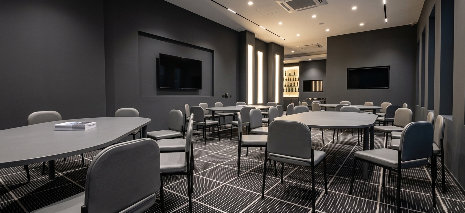An incredibly lifelike, richly detailed, photorealistic wide-angle interior perspective shot of a modern coworking and seminar space set in a sleek, contemporary urban office hub. The scene is illuminated by recessed warm LED ceiling lights and spotlighting that brightly illuminates each table, complemented by the soft, ambient glow of the backlit showcase and screens, creating a sophisticated and business-oriented atmosphere. Captured with a 24mm f/4 tilt-shift lens, emphasizing the rich charcoal matte of the anthracite walls, the fine texture of the black-and-white dotted carpet, and the polished grey surfaces of the furniture, the expansive layout exudes contemporary elegance. Across the room, several large, oval-shaped grey conference tables paired with matching ergonomic grey office chairs are neatly arranged under direct, warm spotlights. Above, the clean white ceiling houses integrated modern linear lighting and minimalist air conditioning vents, contrasting beautifully with the deep anthracite-colored walls. Large television screens are mounted flush on the dark walls, their blank display panels waiting for presentations. Underfoot, the luxurious black carpet with minute white dots adds a sophisticated tactile texture to the floor. In the background, a sleek display cabinet features a brilliantly glowing backlit back panel, casting a soft, warm ambiance that showcases neat rows of bottles on glass shelves, adding a touch of premium hospitality to this professional workspace.