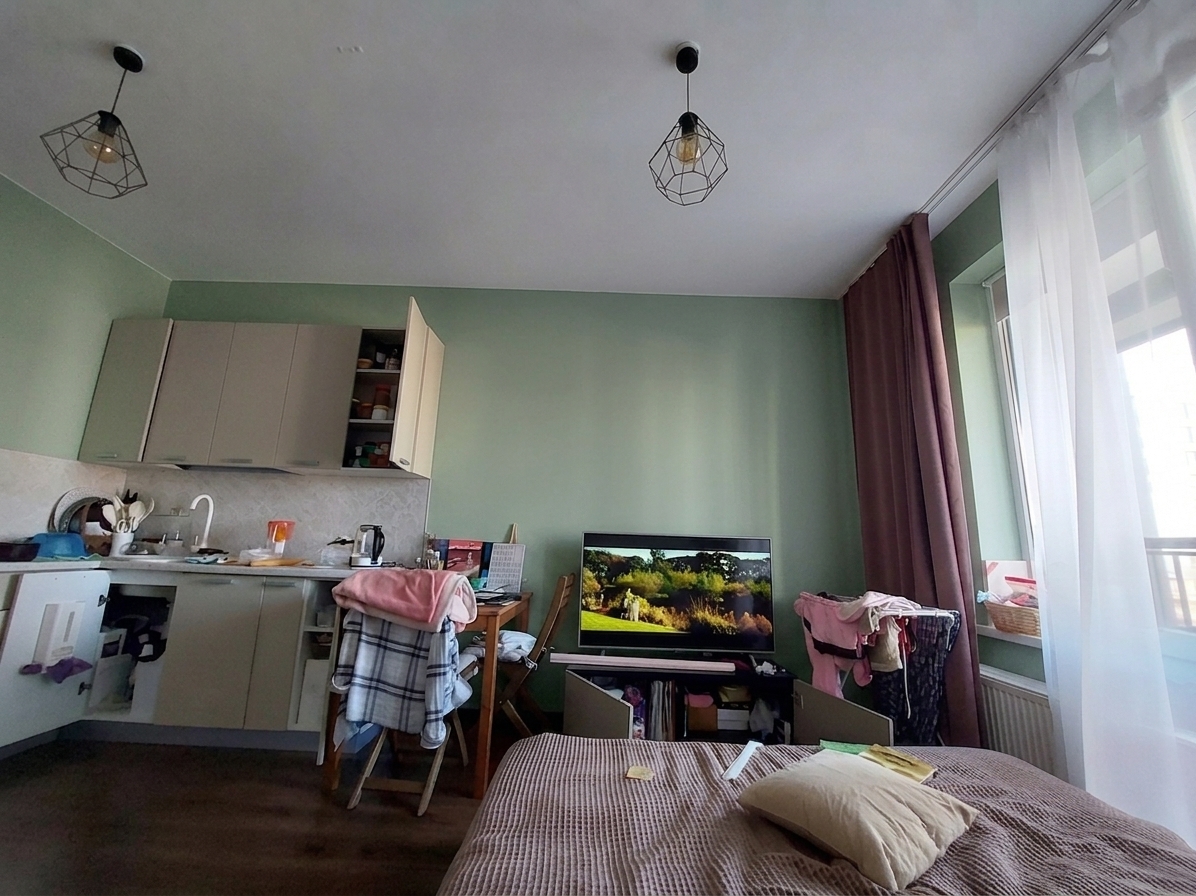 A wide-angle interior photograph of a cozy, lived-in studio apartment, preserving the exact layout, perspective, and composition of the original scene. The previous beige walls have been repainted in a sophisticated, muted dusty sage green with a soft matte finish, showing subtle micro-textures of a paint roller under the natural light. Soft, diffused daylight enters from the window on the right, filtered through the sheer white curtains and casting a gentle gradient of light and shadow across the room. All elements remain perfectly in place: the textured ribbed brown bedspread and soft pillow in the foreground, the casual clutter of clothes draped over chairs and the drying rack, the compact kitchen area with its semi-gloss cabinets and open doors on the left, and the TV screen displaying a colorful outdoor scene. The two wire-frame pendant lights hang from the ceiling with realistic metallic textures. The final image exhibits the authentic characteristics of a high-quality photograph, including a natural depth of field, gentle lens vignetting, soft light bloom near the window, and subtle, realistic shadows in the room's corners.