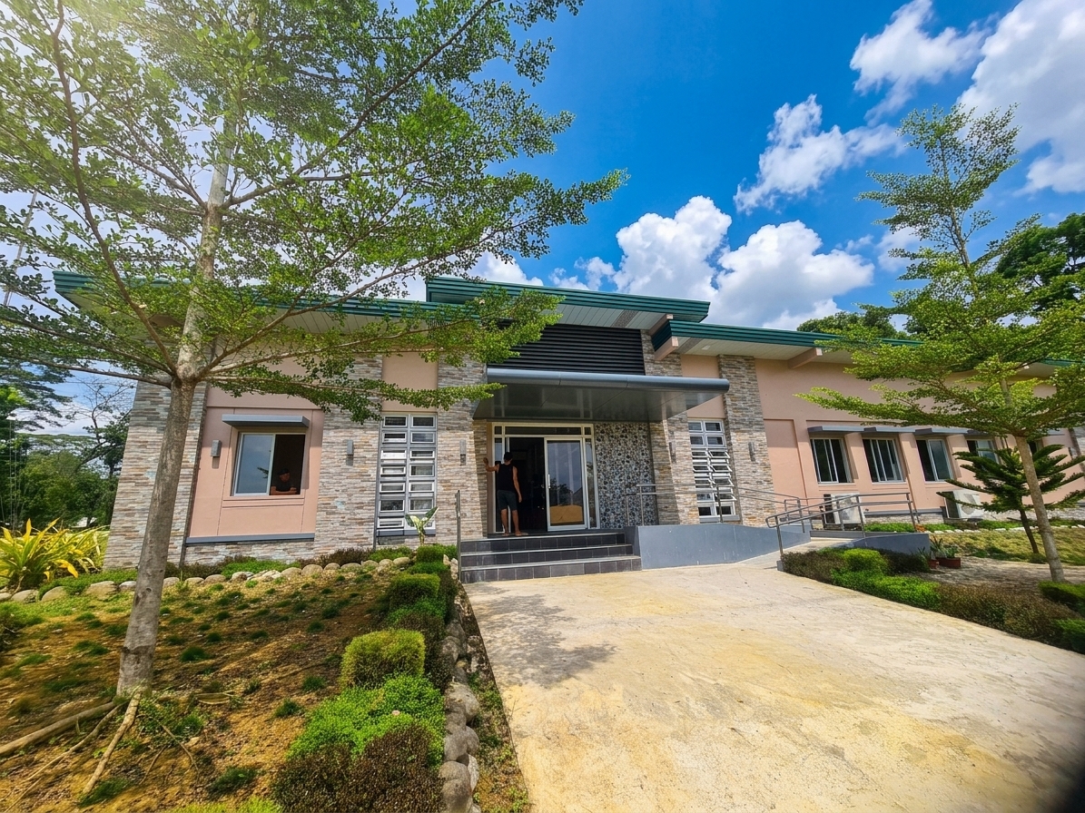 A wide-angle architectural photograph capturing the single-story modern building during a bright, clear spring midday. Crisp, high-overhead sunlight casts sharp, short shadows beneath the dark roof overhang, entrance canopy, and lush foreground trees, illuminating the scene with a bright, natural 5500K daylight color balance. The sky is a vivid spring blue filled with soft, volumetric cumulus clouds that subtly filter the overhead glare. The concrete walkway displays realistic surface texture, fine grain, faint dust, and subtle sun-baked tonal variations leading up to the dark stone entry steps. The exterior features textured stone cladding with matte, multi-tonal slate finishes alongside smooth, warm beige stucco walls showing delicate surface imperfections. Leaves on the surrounding greenery and trees show fresh spring vibrancy, with sunlight glowing through translucent green leaves and casting intricate, dappled light patterns onto the landscaped ground. The glass windows and open glass entry doors feature subtle reflections of the outdoor scene, interior depth shadows, and realistic faint dust smudges on the panes. The photograph exhibits natural depth of field with sharp structural details throughout, gentle lens vignetting at the corners, subtle chromatic aberration along tree foliage against the sky, and a fine, soft film grain.