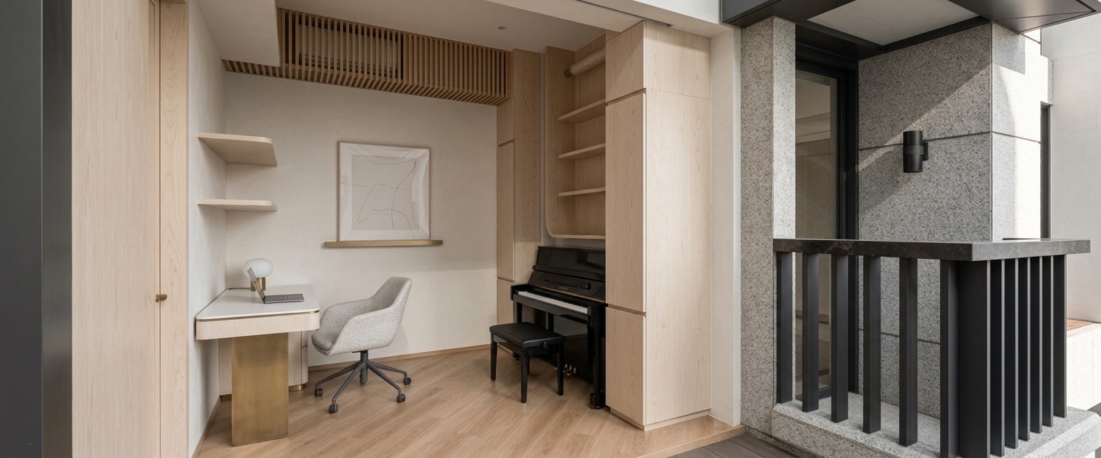 A high-end architectural photograph capturing the exact room layout and composition, illuminated by soft, natural daylight filtering through the right opening. The interior features light birch wood wall paneling with delicate visible grain, matte off-white plaster walls, and warm light-oak hardwood floor planks showing realistic surface texture and soft sheen reflections. On the left, a sleek floating desk with a brushed brass pedestal base and a smooth white laminate top holds a open slim laptop and a small desk lamp, paired with an upholstered light-grey desk chair with detailed fabric weave. Above, a framed minimalist line art piece hangs flat against the subtle wall seam. In the alcove, a satin-black upright piano with matching bench displays subtle reflections along its polished finish under custom built-in light-wood shelving and a slatted timber ceiling soffit. To the right, a textured grey granite exterior column and dark stone balcony counter are rendered with tactile, unpolished stone roughness alongside a dark metal slatted railing and a black wall sconce. The scene is shot on a professional tilt-shift lens maintaining perfectly straight vertical lines, with soft ambient occlusion in the corners, gentle light diffusion across surfaces, natural depth of field, and subtle architectural photography imperfections like faint lens vignetting and fine sensor grain.
