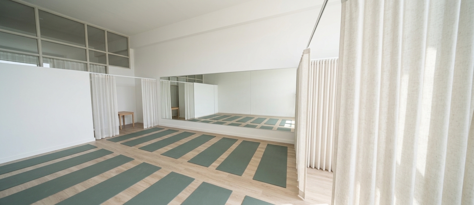 An incredibly lifelike, richly detailed, photorealistic wide-angle interior photo of a yoga studio set in a minimalist fitness haven. The scene is illuminated by bright, soft midday sunlight streaming in on a fresh spring day, creating a serene and invigorating atmosphere. Captured with a professional 24mm architectural lens, emphasizing the subtle grain of light-colored SPC wood flooring, the gentle drape of semi-sheer white curtains framing the side alcoves, and the matte, tactile texture of sage-green yoga mats (in hex #708784) meticulously aligned across the floor. Dominated by pristine white walls, the main wall features a magnificent, large framed mirror that crisply reflects the open space and amplifies the radiant ambient light, offering a calm, harmonious sanctuary tailored for wellness and mindfulness.