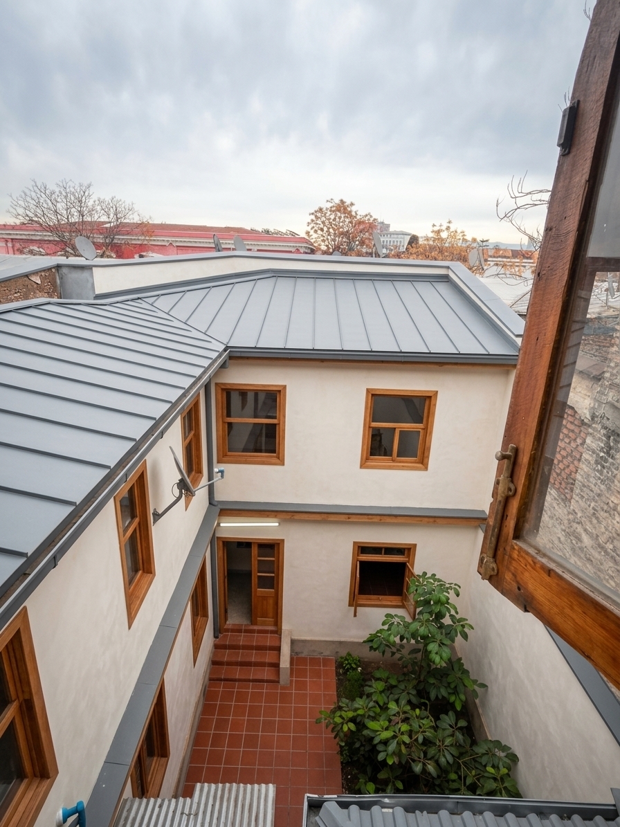 A high-angle architectural photograph looking down into an inner courtyard through an open upper window, keeping the exact composition, geometry, perspective, and framing of the original image. The roofs are fully updated with smooth matte ash gray standing-seam zinc panels, displaying clean seam lines and a muted RAL 7040 finish. The surrounding exterior courtyard walls are freshly coated in a soft ivory shade featuring a chalky, lime-washed plaster texture with natural micro-surface imperfections. All door and window frames are crafted from rich natural wood with visible timber grain and a warm satin finish. The lower courtyard floor is paved with traditional Chilean colonial red clay tiles in a rich terracotta hue, laid in a clean grid layout. The lush green foliage in the courtyard remains intact in its exact original placement. The scene is illuminated by soft, overcast natural daylight from the cloudy sky above, casting smooth ambient shadows and highlighting the tactile quality of the materials, captured with a sharp wide-angle lens, deep depth of field, delicate vignetting, and natural photographic grain.