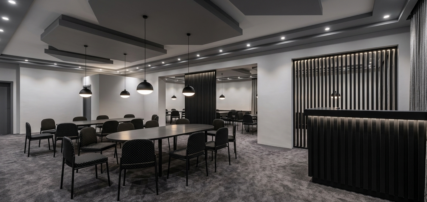 An incredibly lifelike, richly detailed, photorealistic wide-angle interior photograph of a modern fusion meeting room set in an upscale contemporary commercial space. The scene is illuminated by soft ambient illumination from matte white recessed spotlights along the ceiling and warm downward pools of light from hanging dome pendants, complemented by dramatic LED strip backlighting nestled between vertical slats, creating a sophisticated, refined, and moody atmosphere. Captured with a 24mm tilt-shift architectural lens, emphasizing the plush textures of the dark gray carpet, polished charcoal surfaces, and crisp geometric lines. Plush dark gray carpet covers the expansive floor, providing a soft foundation for sleek dark gray dining and conference tables surrounded by elegant black upholstered chairs with fine white speckled dots. The multi-level ceiling features stepped geometric tiers in complementary shades of gray, contrasting against smooth, light gray surrounding walls. Overhanging dome pendant lights are styled with matte black exteriors and luminous white inner bowls, casting targeted light onto the seating below. To the right, a prominent reception desk clad in vertical black slats with ambient backlighting echoes the tall slatted partition screens that gracefully divide the spacious interior, adding architectural depth, exquisite texture, and a high-end designer aesthetic.