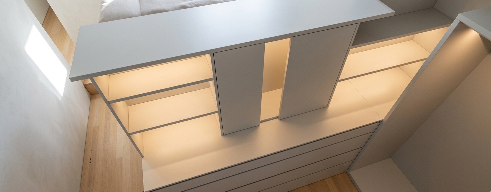 A high-angle architectural interior photograph of a modern minimalist dressing closet, faithfully maintaining the exact layout, geometry, framing, and camera angle of the scene. The custom built-in cabinetry is rendered with ultra-realistic matte off-white and light gray satin lacquered wood panels featuring subtle surface micro-textures, precise panel seams, and realistic drawer reveals. Warm, soft LED strip lighting with a cozy 2800K color temperature is seamlessly integrated beneath shelves and within recessed compartments, casting a gentle glow that highlights the interior structure. Light blonde oak hardwood flooring with fine natural grain and a soft satin finish spans the visible floor space. The illumination is bright and airy, blending the warm ambient LED accents with soft, diffused daylight pouring in from the side, creating soft directional shadows and realistic light bounce throughout the alcoves. Captured with a sharp wide-angle lens, the image displays clean photographic realism with faint camera grain, delicate specular highlights along panel edges, and true-to-life material physics without altering any structural elements.
