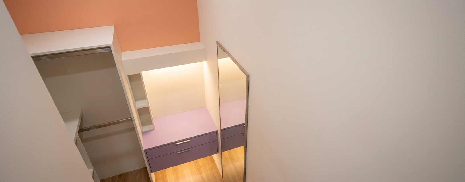 A high-angle interior photograph of a modern minimalist walk-in closet, perfectly maintaining the exact geometry, structural layout, camera angle, and furniture design from the image. The built-in vanity unit features smooth, matte lavender-colored drawer fronts with sleek brushed-metal handles, topped with soft, warm LED strip lighting hidden beneath the white upper shelving and soffit, casting an inviting 2800K glow down onto the surfaces. The upper wall section is painted in a warm, subtle peach-coral shade, contrasting gracefully with the clean off-white painted walls. The floor is made of realistic light oak wood planks with natural wood grain and a soft satin finish that gently catches the ambient light. On the right, the framed full-length mirror reflects realistic soft shadows and warm ambient illumination. Shot with a professional camera using a wide-angle lens at f/4, the photograph features realistic material textures including micro-imperfections on painted surfaces, subtle contact shadows under the floating drawers, gentle lens vignetting near the frame edges, and fine, natural grain that brings life to the architectural space.