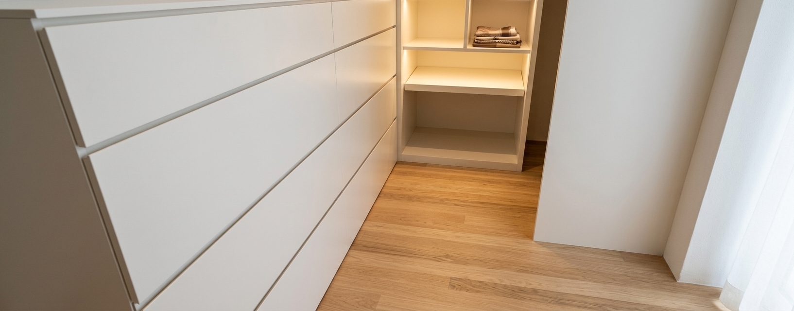 A high-end architectural photograph of a modern minimalist walk-in closet in bright, airy light tones, preserving the exact camera angle, composition, and layout of the scene. The sleek handleless drawer cabinet on the left is finished in smooth satin off-white matte lacquer, showing subtle micro-textures and soft ambient reflections along its clean horizontal recessed channels. The inset alcove unit directly ahead serves as an elegant modern shoe rack, warmly illuminated from within by concealed 3000K recessed LED strip lights that cast a soft downward glow and highlight its interior shelving structure. The floor is clad in light oak wood planks with authentic grain variation, soft satin sheen, and faint seam details that stretch along the perspective lines. Ambient warm light fills the room, creating realistic soft contact shadows where the furniture meets the floor and walls. Shot with a sharp architectural 24mm lens, the image displays lifelike material properties, precise edge highlights, balanced exposure with smooth highlight roll-off, and subtle natural photographic qualities like faint lens vignetting and micro-grain.