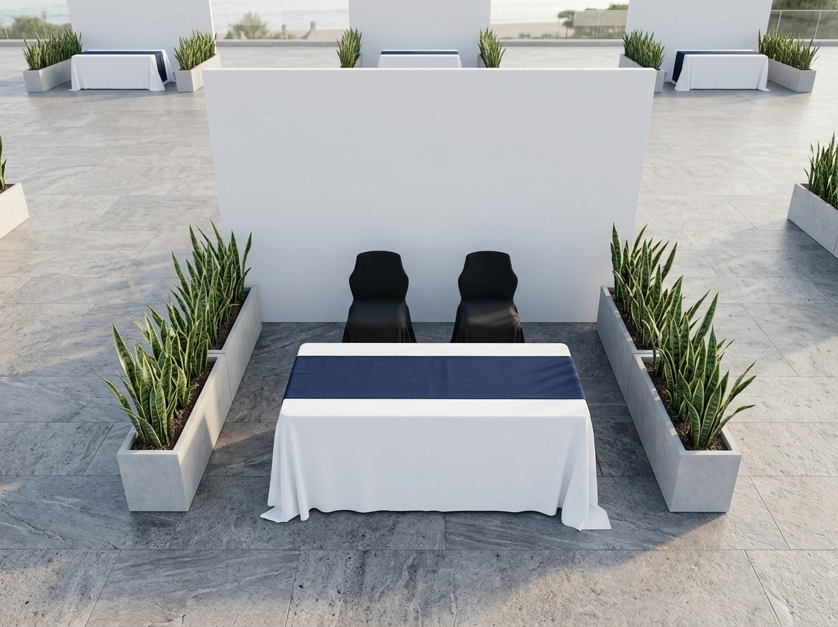 An incredibly lifelike, richly detailed, photorealistic top-down architectural rendering of a contemporary event booth module set in an open-air coastal patio venue. The scene is illuminated by radiant, soft coastal daylight and gentle ambient shadows carried by an ocean breeze, creating an airy, sophisticated, and inviting atmosphere. Captured with an overhead medium-format camera, emphasizing realistic fabric draping, lush foliage textures, and refined material finishes. At the center, the rectangular display table is dressed in a crisp, flowing white tablecloth layered with an elegant, deep navy blue runner. Two modern, matte black ergonomic chairs tuck neatly behind the table against a smooth, minimalist white back wall partition. Flanking either side, elongated light gray concrete planter boxes are lushly planted with upright, architectural Sansevieria (snake plants), extending to serve as natural green privacy dividers between adjacent, identical exhibition stands across the sleek stone patio.