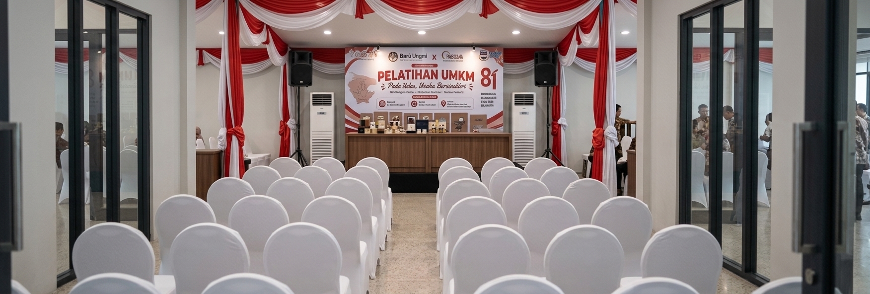 An incredibly lifelike, richly detailed, photorealistic wide-angle interior shot of a formal event and seminar hall set in a contemporary multipurpose venue. The scene is illuminated by soft, diffuse ambient interior lighting seamlessly blended with crisp stage spotlights that accentuate the presentation platform, creating a professional, celebratory, and engaging atmosphere. Captured with a wide-angle 24mm architectural lens at eye level with refined depth of field, emphasizing the delicate silk textures of the draped red-and-white ceiling swags, the smooth matte fabric slipcovers on the curved banquet seating, and the rich wood-grain paneling of the main presentation counter. Framed symmetrically through sleek charcoal door jambs, the perspective leads past neatly arranged rows of pristine white chairs toward the brightly lit stage, equipped with high-fidelity PA speakers, modern standing AC units, and a sharp, high-resolution backdrop banner complete with an elegant product display arrangement.