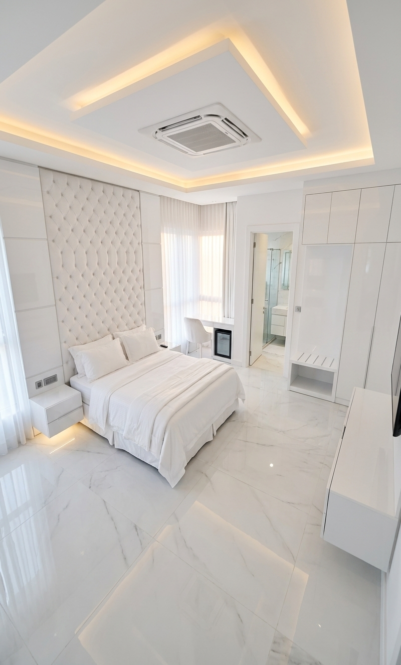 An incredibly lifelike, richly detailed, photorealistic high-angle architectural photograph of a bespoke luxury hotel suite set in a modern minimalist haven. The scene is illuminated by the soft, warm golden glow of an early autumn sunrise filtering gracefully through sheer white floor-to-ceiling draperies, balanced by warm, indirect LED cove lighting recessed within the geometric false ceiling, creating a serene, ethereal, and sophisticated atmosphere. Captured with a professional medium-format camera and a wide-angle architectural tilt-shift lens, emphasizing the interplay of brilliant high-gloss reflections, deep velvet textures, and flawless monochromatic craftsmanship.

The suite unfolds in an immaculate palette of pure, luminous whites. The floor is composed of large-format, mirror-polished white marble tiles that catch the morning light and cast crystal-clear reflections of the entire room's furnishings. Centered on the left, a king-sized master bed is dressed in layered, crisp white Egyptian cotton linens, a folded white duvet, and plump sleeping pillows. At its head stands a magnificent full-height feature wall boasting a white velvet diamond-cut tufted headboard, framed by glossy white lacquered architectural panels with fine vertical bead moldings. Flanking the bed are floating, glossy white nightstands equipped with sleek integrated controls and subtle under-cabinet ambient illumination.

Suspended overhead, the crisp white multi-tiered tray ceiling incorporates a clean, flush-mounted centralized cassette air conditioning unit, surrounded by subtle perimeter cove lighting that washes the ceiling in a gentle, warm halo. Near the entryway, the storage area features an integrated glossy white luggage rack with protective chrome ribs, transitioning smoothly into a tailored, floor-to-ceiling wardrobe with flush-fit seamless white doors. 

Opposite the bed, a minimalist white floating entertainment console neatly conceals a mini-fridge cabinet beneath a pristine wall setup, leading to the bright corner window area where a sleek white lacquer study desk rests beside a plush, ergonomically sculpted white velvet chair. Through a tailored white doorway at the upper quadrant, the ensuite bathroom is visible, finished in bright white stone finishes, a frameless glass walk-in shower enclosure, and a modern white designer vanity. The entire composition radiates pristine tranquility, where every texture—from matte velvet to high-gloss lacquer—creates subtle depth and elegance within the all-white sanctuary.