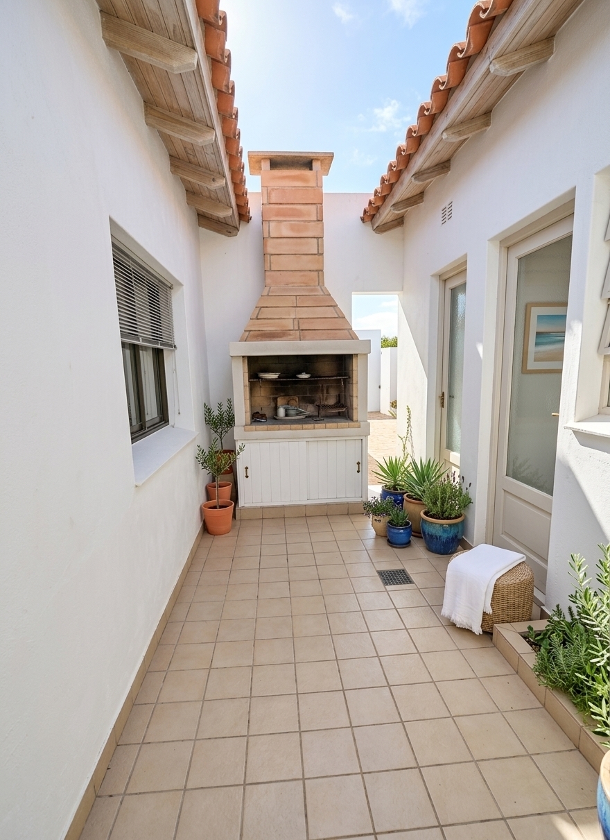 An incredibly lifelike, richly detailed, photorealistic wide-angle architectural shot of a revitalized outdoor patio and courtyard set in a sun-drenched seaside home. The scene is illuminated by bright, sun-kissed natural daylight pouring gently between the terracotta-tiled roof eaves, creating an airy, light, and wonderfully relaxed nautical atmosphere filled with a sense of coastal breeze. Captured with a full-frame camera using a 24mm architectural tilt-shift lens, emphasizing the crisp textures of whitewashed stucco walls, warm terracotta roof tiles with exposed wood rafters, and sandy-neutral paved flooring. Anchoring the far end of the terrace, the tall traditional outdoor fireplace is refreshed with light masonry accents, complemented by serene framed seascape wall art hung along the freshly painted walls. The inviting space is styled with uncluttered simplicity, featuring casual wicker and light natural wooden furniture dressed in soft shades of coastal blue, crisp white, and sandy neutrals, while vibrant potted coastal plants and olive greenery in glazed blue ceramics breathe organic life into this open, beach-inspired retreat.