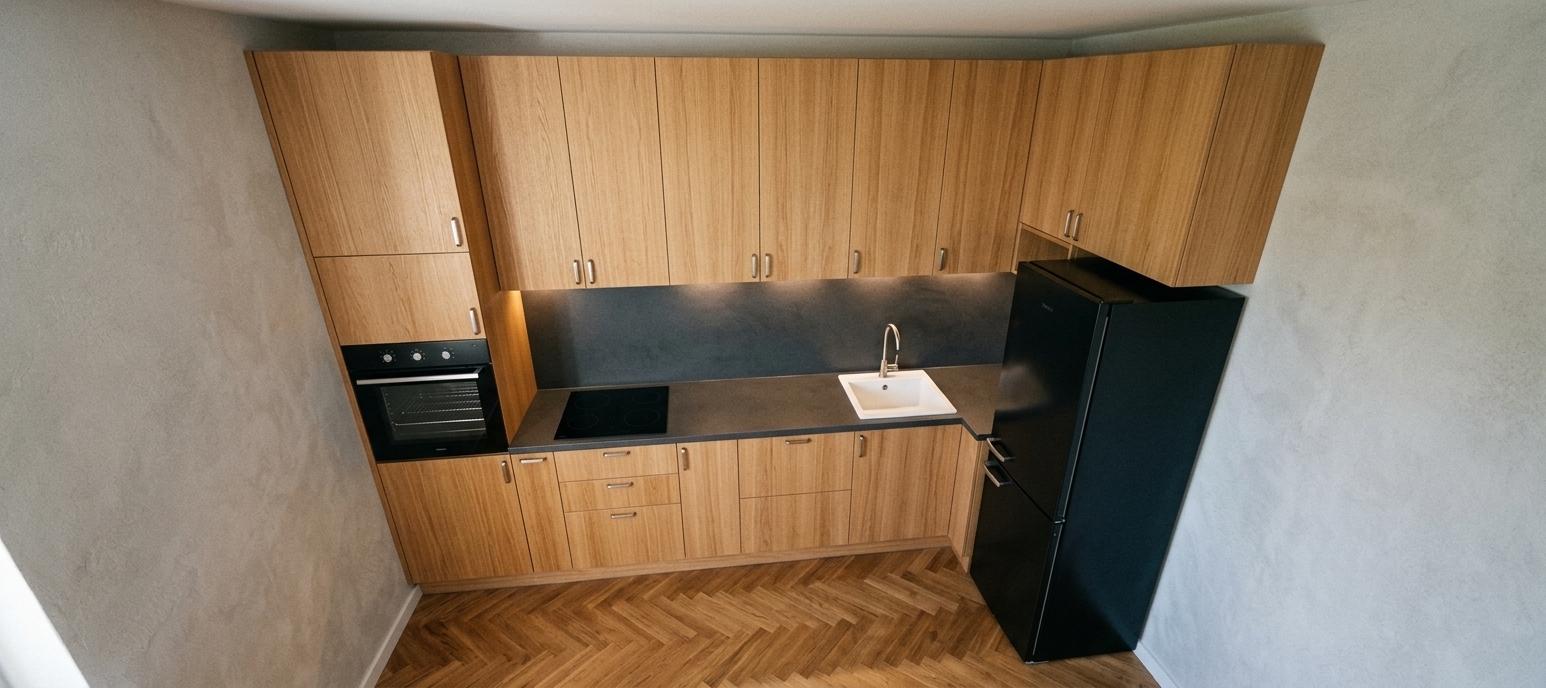 An incredibly lifelike, richly detailed, photorealistic wide-angle interior shot of a modern, streamlined kitchen and coffee station set in a minimalist haven. The scene is illuminated by soft, natural ambient daylight paired with gentle under-cabinet LED illumination, creating a serene, zen-inspired atmosphere. Captured with a professional 35mm architectural lens, emphasizing the warm, vertical grain of the seamless oak cabinetry, the rich matte-black finish of the freestanding refrigerator, and the deep charcoal backsplash and countertops. The clean layout features an integrated built-in oven, a flush black ceramic cooktop, and a crisp white undermount sink set against textured light-gray walls and warm parquet hardwood flooring. Clutter-free surfaces, streamlined silver handles, and balanced proportions evoke an elegant, functional, and tranquil modern sanctuary.