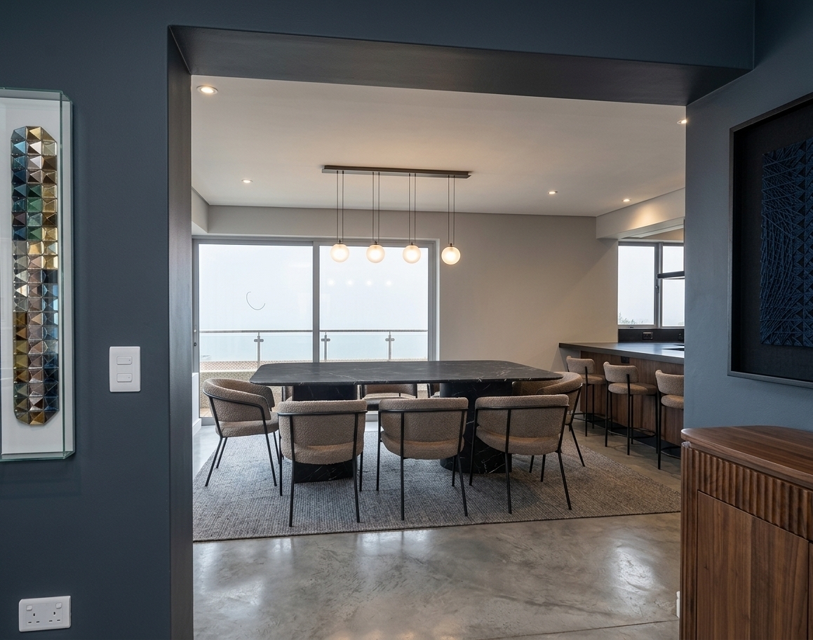 The photo captures a sophisticated dining room, embodying a modern fusion aesthetic with its sleek lines and thoughtful material choices, viewed through a dark, wide archway from an adjacent space. In the foreground, partially visible on the right, is a contemporary wooden console table with a striking fluted texture, providing a warm contrast to the cooler tones of the room. The dining area is anchored by a large, rectangular table with a dark, perhaps polished stone or wood finish, surrounded by six modern dining chairs featuring thin, dark metal frames and light-colored upholstered seats and backs, suggesting comfort and minimalist design. Above the table, a linear pendant light fixture with four spherical, glass globes casts a gentle glow, complemented by several recessed lights in the ceiling, illuminating the space evenly. Beyond the dining table, expansive, floor-to-ceiling windows with slender dark frames dominate the back wall, suggesting ample natural light and offering a glimpse of an outdoor balcony with a minimalist railing, implying an urban or elevated setting. To the right, the dining room seamlessly transitions into a sleek kitchen or bar area, indicated by a dark counter with matching modern bar stools, further emphasizing the open-concept layout. On the left wall, a striking vertical piece of abstract art with colorful, geometric tessellations in a clear frame adds a vibrant, artistic touch, while another framed dark, grid-patterned artwork is visible on the far right wall, contributing to the room's curated and refined atmosphere. The overall impression is one of modern elegance and functional design, where various textures and contemporary furnishings blend harmoniously to create an inviting yet stylish living space.