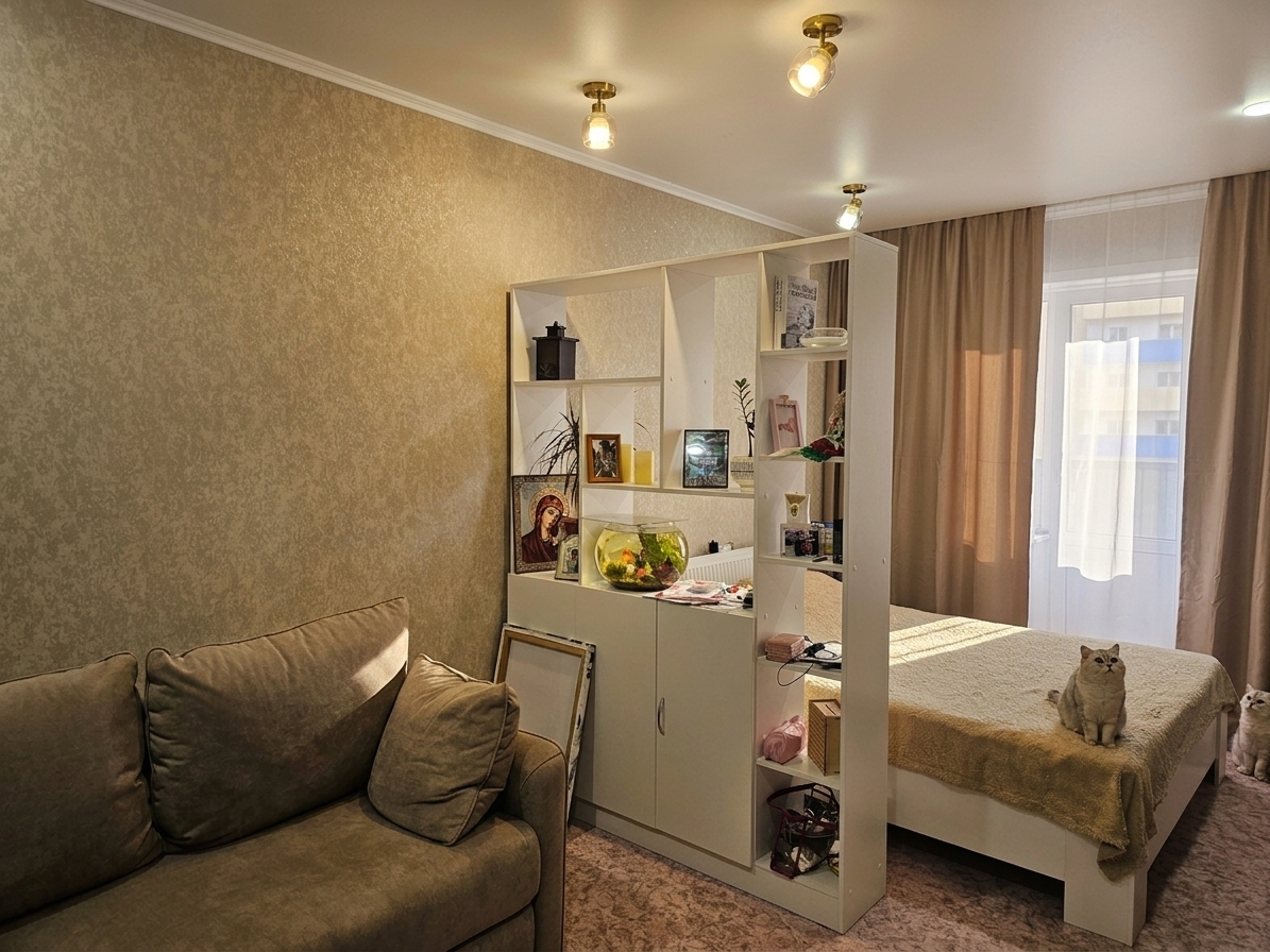A realistic photograph of the exact room interior maintaining its existing layout, geometry, and neutral beige palette, enhanced with vivid lighting accents. Balanced natural light filters softly through the balcony curtains, while warm ceiling spotlights cast defined pools of light and gentle shadows, highlighting the shimmering texture of the wallpaper, the fabric weave of the sofa, and the glass reflections of the fishbowl. Captured with a sharp wide-angle lens, featuring accurate specular highlights, realistic fabric folds, mild depth of field, subtle film grain, and gentle vignetting to create an authentic photographic look.
