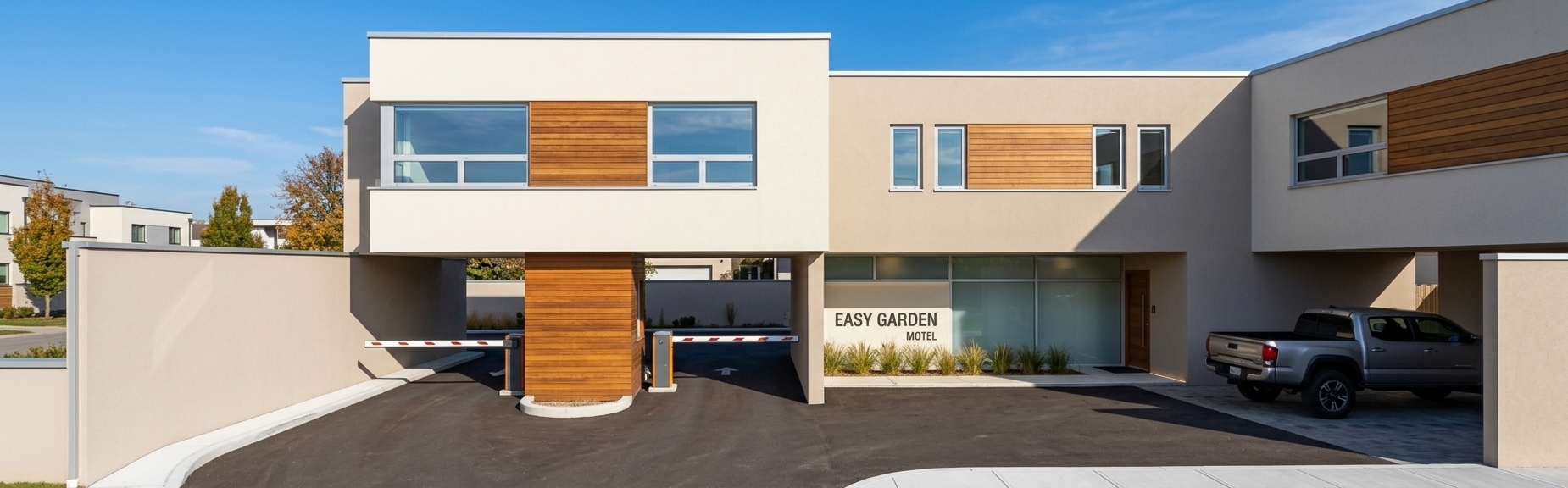 An incredibly lifelike, richly detailed, photorealistic eye-level exterior architectural shot of the modern entrance and minimalist reception of "Easy Garden Motel" set in a calm, contemporary suburban setting. The scene is illuminated by crisp, direct midday autumn sunlight casting sharp, dynamic shadows under the floating cantilevered volumes, creating a serene, elegant, and welcoming minimalist haven. Captured with a professional 24mm tilt-shift architectural lens, emphasizing the rich natural grain of horizontal teak wood cladding panels, smooth off-white and soft beige exterior stucco framed by clean aluminum coping trim, and expansive modern window apertures reflecting a vivid blue sky. At the ground level, a dark asphalt driveway guides vehicles past twin automated barrier gates flanking a warm wood-wrapped security column, while the ground-floor lobby presents luminous frosted glass partitions, subtle integrated LED wall wash lighting, a refined timber entry door, and manicured ornamental grasses resting cleanly beside the covered carport.