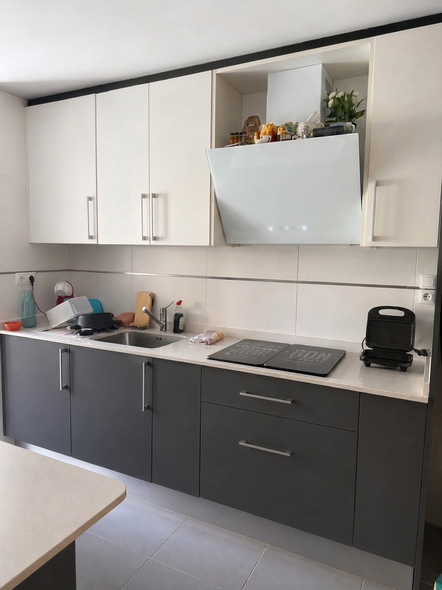 A realistic interior photograph maintaining the exact camera angle, layout, and composition of the kitchen, featuring the existing dual-tone dark grey and cream cabinetry, tiled backsplash, and countertop accessories, with the central range hood replaced by a modern, angled white tempered glass decorative extractor hood. The scene is illuminated by soft, natural daylight entering from the side, producing crisp reflections across the sleek white glass hood, polished chrome handles, and quartz counter. Surface textures show authentic micro-imperfections, realistic specular highlights, subtle contact shadows, and faint natural camera grain with gentle vignetting for a clean, photorealistic finish.