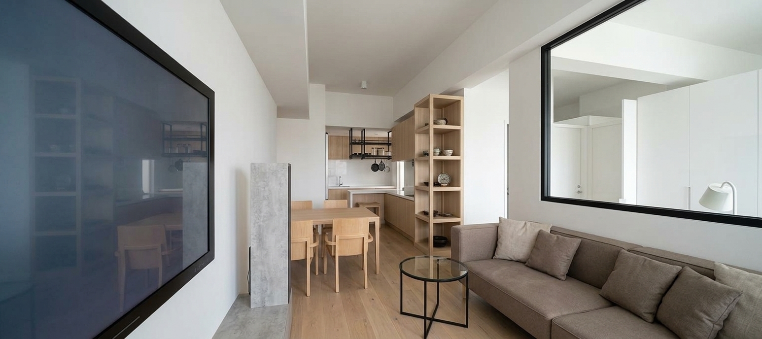 An incredibly lifelike, richly detailed, photorealistic wide-angle interior shot of a bright, modern minimalist living and dining room set in an airy contemporary urban apartment. The scene is illuminated by crisp, pure midday light washing gently across pristine white walls, creating an impeccably calm, luminous, and refined minimalist haven atmosphere. Captured with an architectural full-frame camera and a wide-angle tilt-shift lens, emphasizing the seamless interplay of organic light wood textures, smooth matte surfaces, and clean architectural lines. On the right, a low-slung, tailored taupe sectional adorned with contrast throw pillows sits beneath a sleek black-framed interior window opening into an adjacent room, beside a minimalist circular accent table resting on warm, wide-plank oak flooring. To the left, a massive flush-mounted smart display anchors the wall above a textured stone-finish console, while the linear perspective naturally pulls the eye toward the dining ensemble and an open kitchen finished with light blonde millwork, quartz countertops, and a chic suspended black metal rack.
