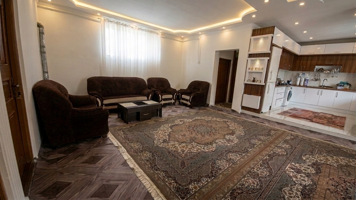 A wide-angle interior photograph of the living room and open kitchen, featuring a timeless, clean plaster ceiling with softly diffused warm cove lighting and subtle recessed spotlights. Gentle daylight filters through the sheer white window drapes, balancing with the warm ceiling ambient glow to cast soft, natural shadows across the space. The plush textures of the brown armchairs, the detailed patterns of the large Persian rugs, and the wooden textures of the flooring and kitchen cabinetry are rendered with natural surface imperfections, authentic depth, and faint photographic grain.