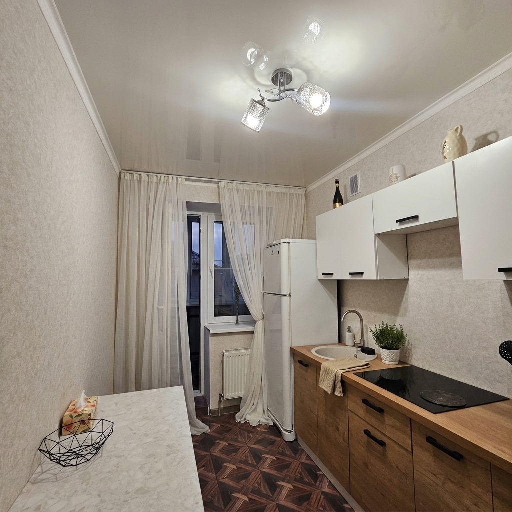 A photorealistic wide-angle interior photograph preserving the exact room layout, framing the far window with two light, semi-sheer white tulle curtains gracefully draped on both sides. Maintain the identical cabinetry, patterned flooring, table, and structure while adding subtle cozy decor accents such as a small potted herb and folded linen near the sink. The scene is illuminated by warm ambient light from the existing ceiling fixture mixed with cool evening light through the glass door, capturing delicate fabric translucency, realistic surface textures, soft natural shadows, gentle lens vignetting, and fine photographic grain.
