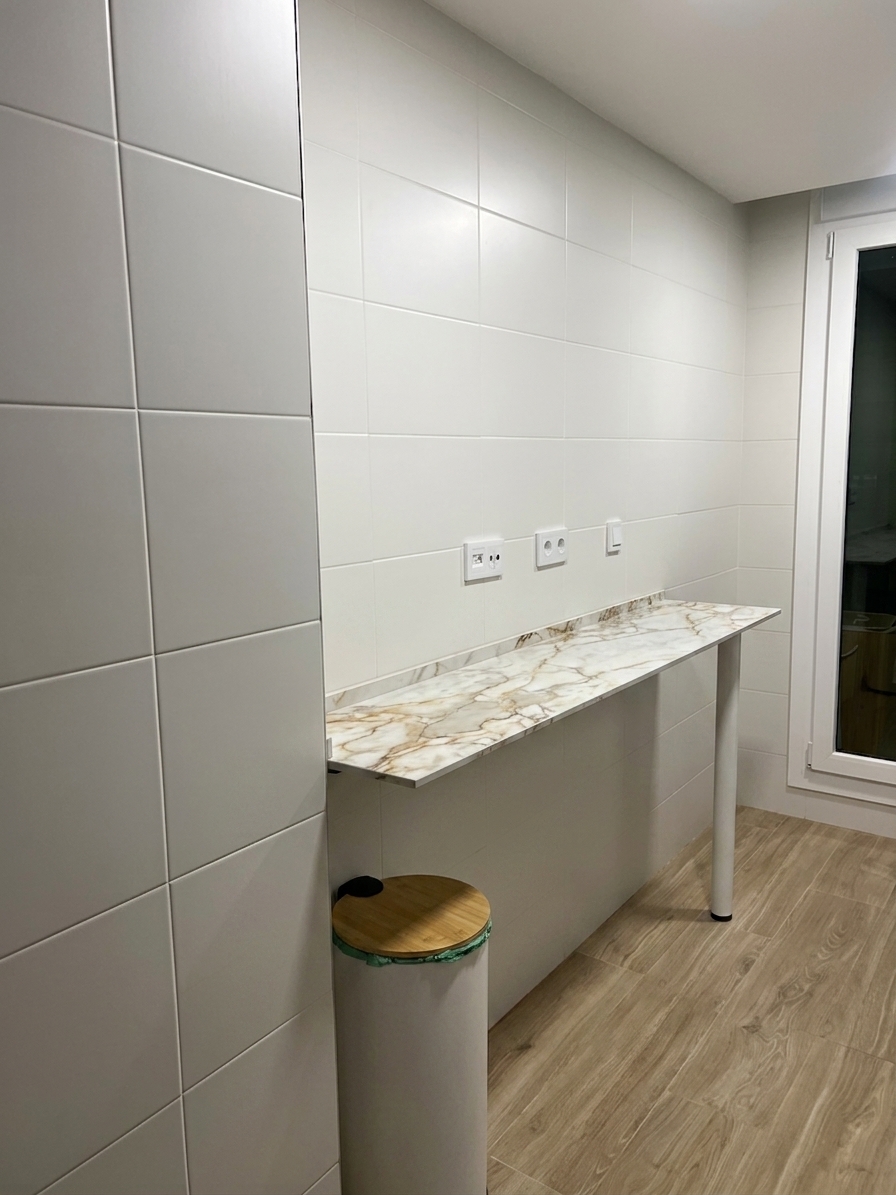 A sharp architectural photograph of the kitchen space, seamlessly incorporating a slim 12mm-thick Laminam Oro del Jerte porcelain bar countertop measuring 175cm wide by 39cm deep mounted directly beneath the wall outlets, supported on its right end by a 110cm tall round white metal peninsula leg. The scene retains the exact camera position, wall tile grid, and wood-look flooring, illuminated by soft diffuse indoor light that brings out the delicate warm veining and satin sheen of the stone slab. Subtle contact shadows anchor the leg to the floor, captured with a standard lens perspective, crisp focus, balanced neutral color grading, and realistic photographic depth.