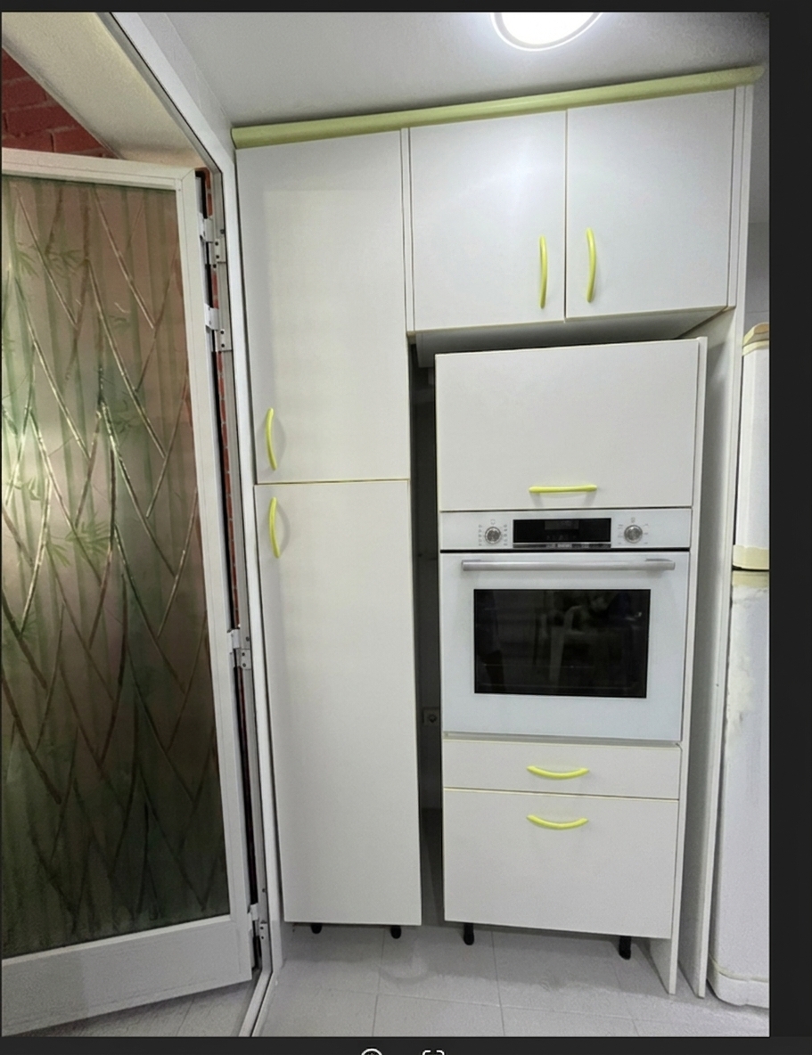 A realistic photograph of the kitchen cabinetry maintaining the exact framing and perspective, featuring finished white laminate cabinetry with matching light lime-green curved handles, enclosing the open compartments with seamless matching white cabinet fronts. Soft, diffused natural indoor lighting illuminates the matte white surfaces, casting subtle realistic shadows and authentic reflections on the oven glass panel and patterned translucent glass door to the left. Fine photographic details include natural micro-textures on the melamine finishes, crisp seam lines between cabinets, gentle ambient occlusion, and subtle natural lens falloff with authentic photographic grain.