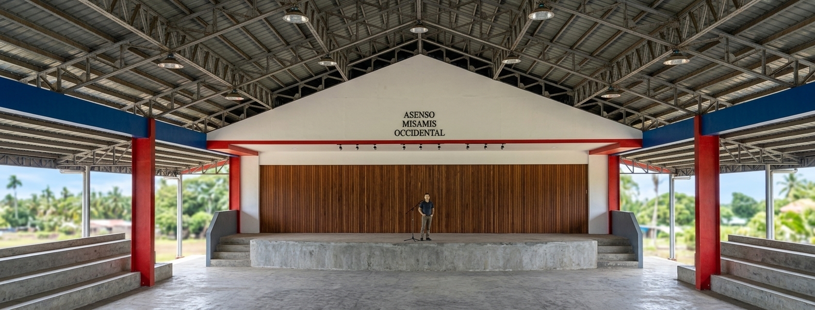 A bright architectural photograph of the open-air covered pavilion captured under harsh midday summer sunlight, casting crisp overhead shadows beneath the high roof structure. The complex exposed steel trusses and corrugated metal ceiling feature realistic industrial textures, while the painted red pillars and blue trim show natural semi-gloss finishes with subtle weathering. The white gabled pediment clearly displays the black lettering reading ASENSO MISAMIS OCCIDENTAL above the concrete stage and vertically slatted wood backdrop. The concrete bleachers and flooring exhibit authentic micro-textures, fine dust, and light scuffs, with a real person standing naturally at center stage. Shot on a wide-angle lens with balanced dynamic range, clear highlights, and sharp architectural detail throughout.