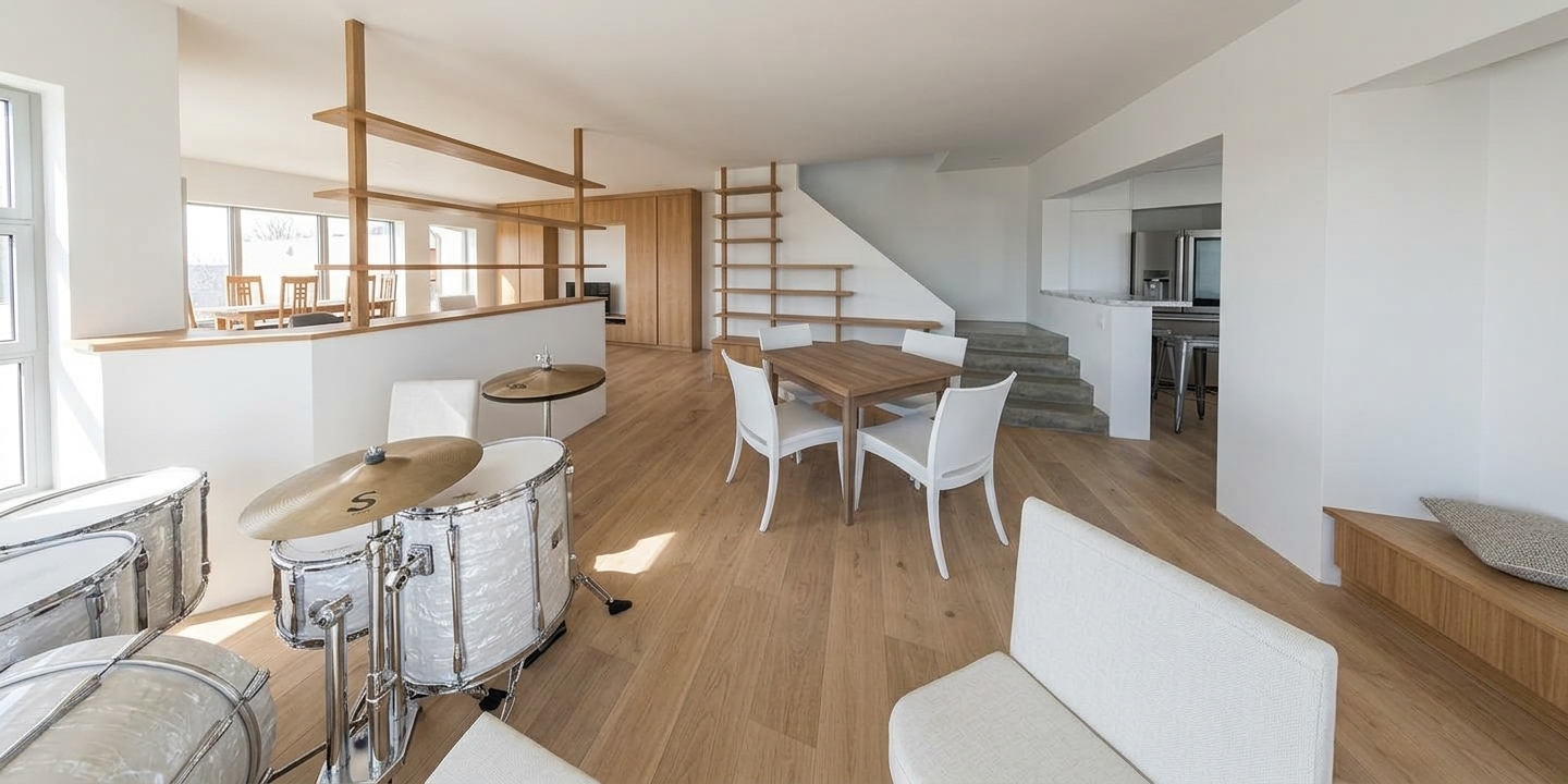 A wide-angle interior architectural photograph bathed in bright, neutral midday sunlight streaming through the rear windows. The space features smooth white painted walls, natural pale wood plank flooring, and warm timber shelving. In the foreground, a drum kit displays realistic gloss lacquer finishes, brushed brass cymbals, and polished chrome hardware with crisp highlights. The central dining table reveals a natural matte wood grain surrounded by contemporary white chairs casting soft, defined midday shadows. Clear, high-key ambient illumination fills the multi-level room, bringing out authentic surface textures, subtle wall paint sheen, and realistic material depth across the stairs and open kitchen view with neutral color temperature and clean optical clarity.